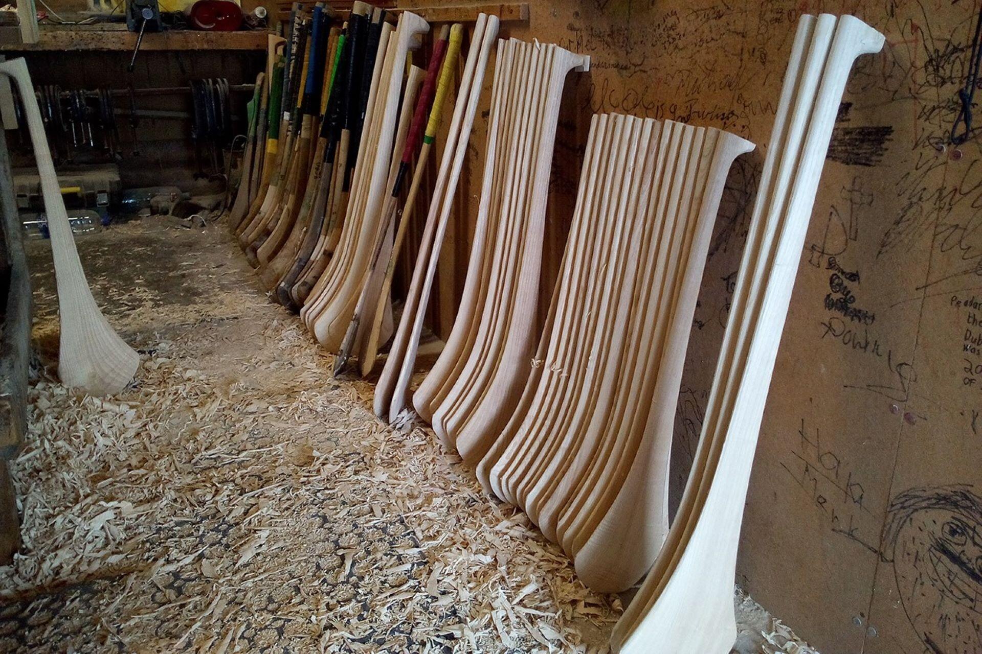 Rows of finished and unfinished wooden hurleys standing in a workshop beside wood shavings.