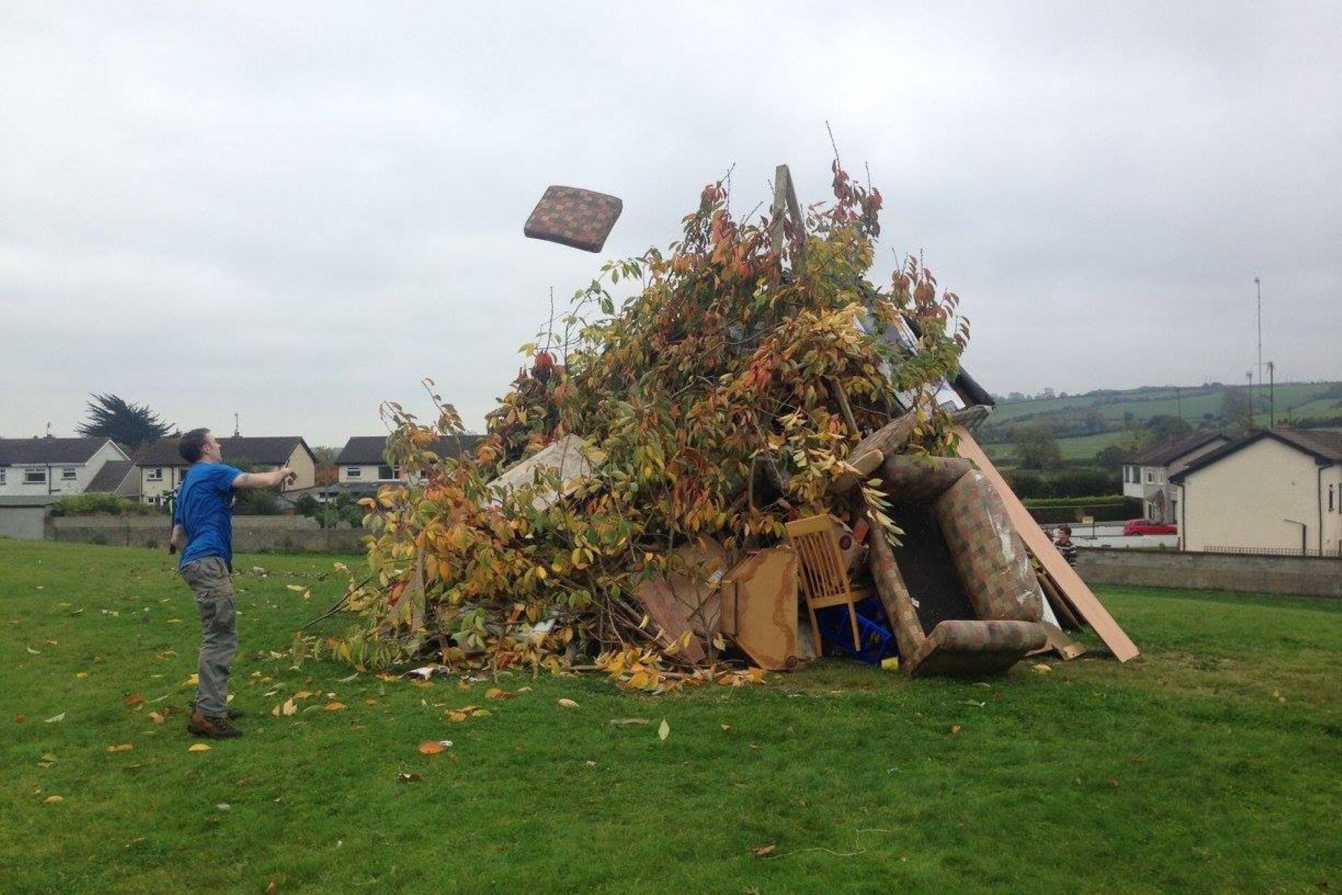 Man throwing a mattress onto a large bonfire pile on a grassy field