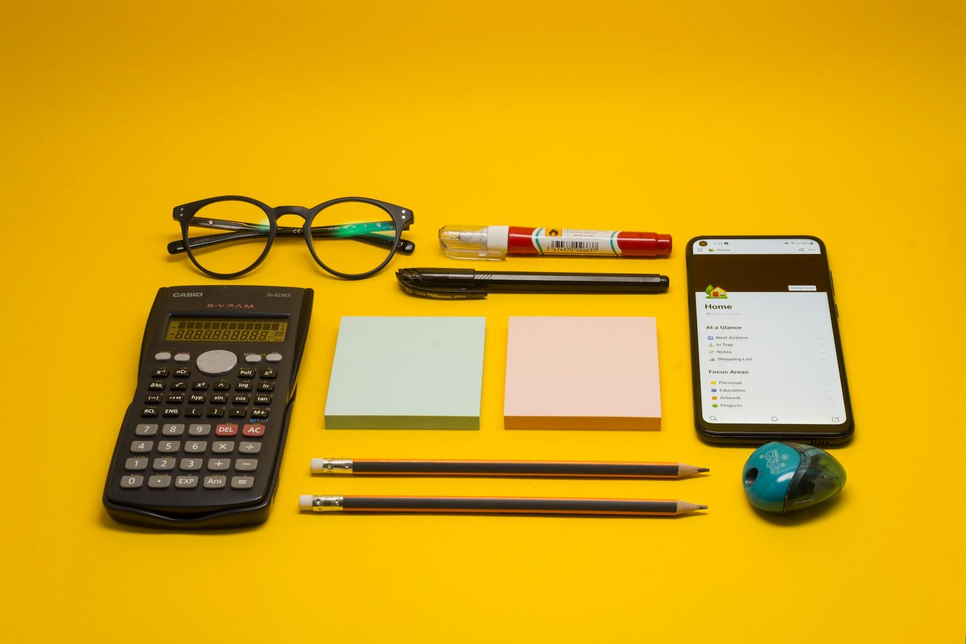 Calculator, glasses, pencils, sticky notes and a smartphone arranged on a yellow surface
