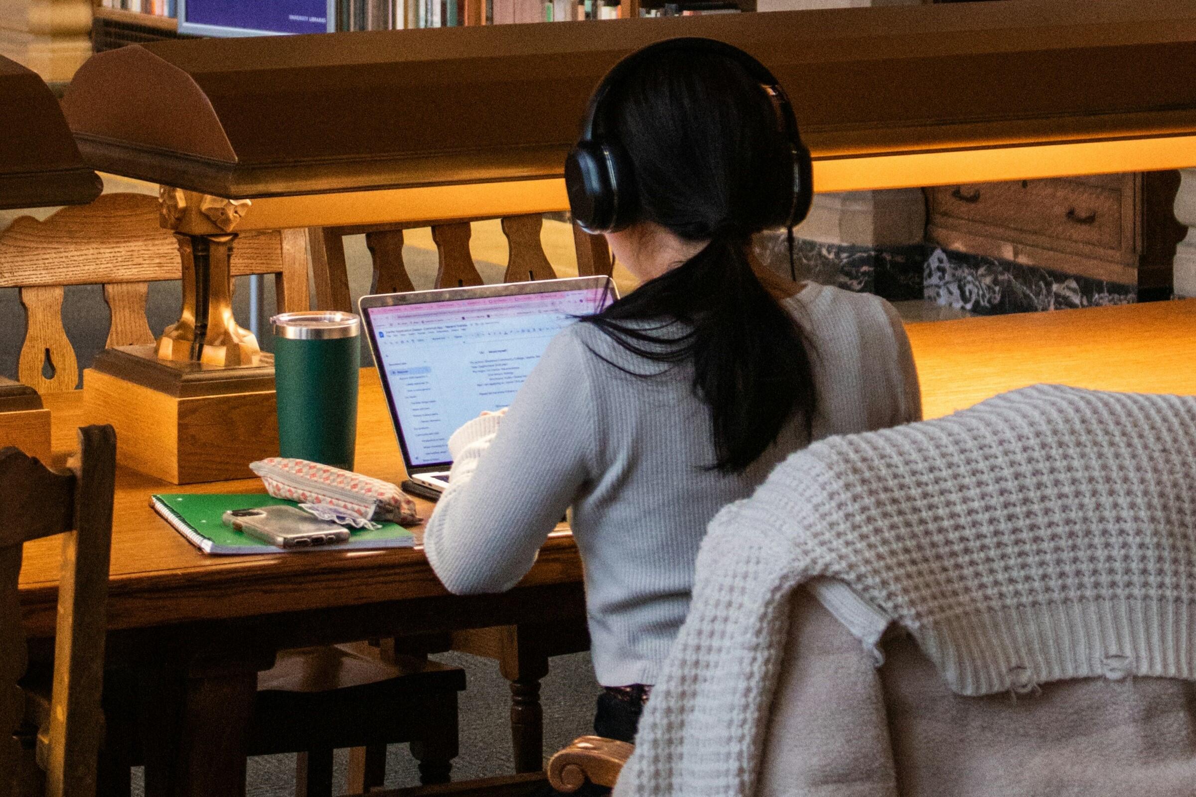 Student using a laptop at a library desk while wearing headphones