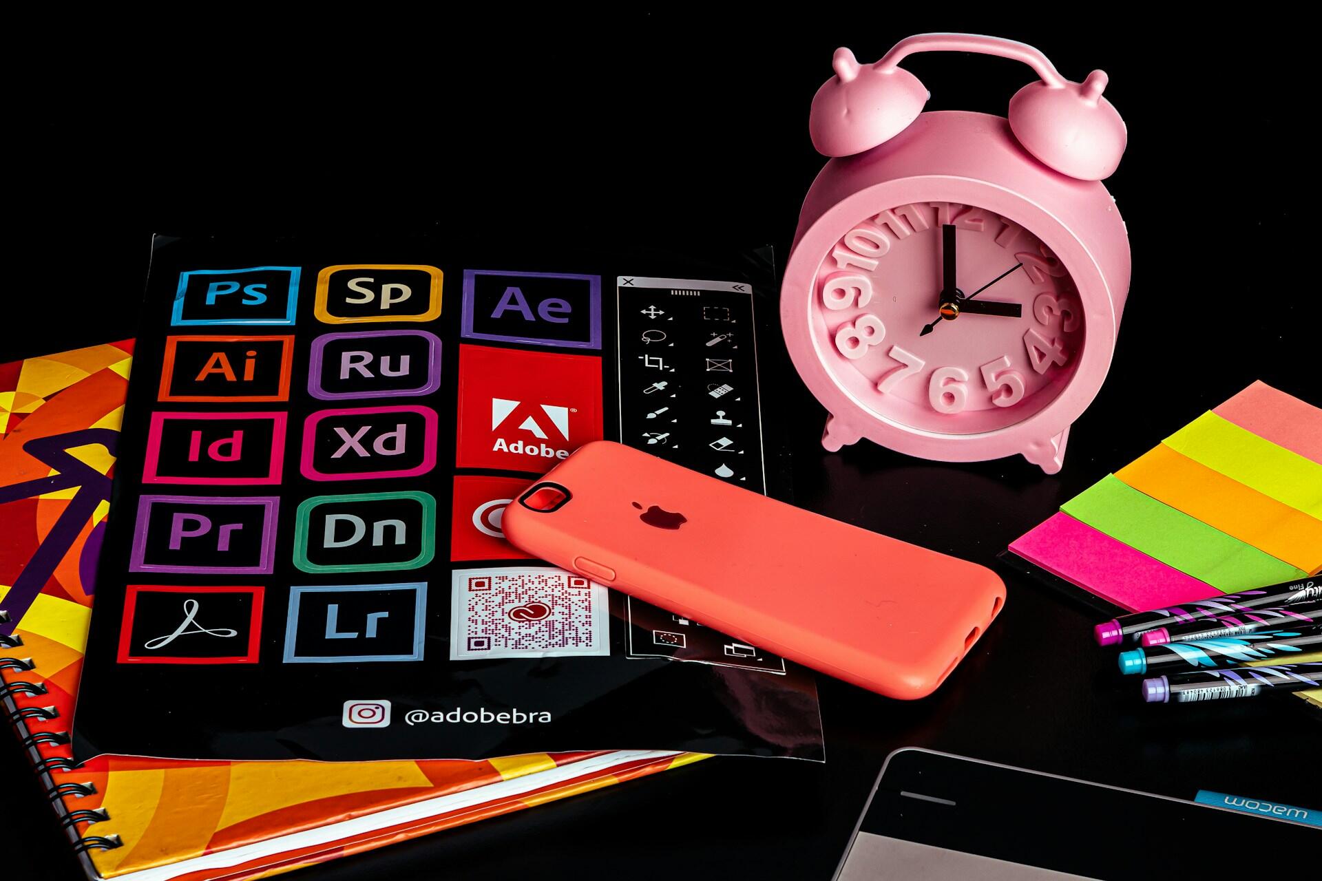 Pink alarm clock beside a phone, notebooks, sticky notes and stationery