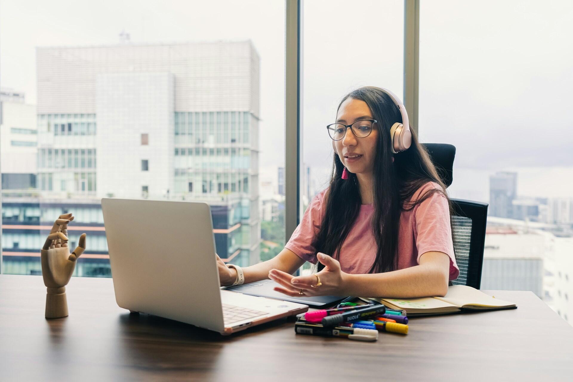 Woman wearing headphones speaking at a laptop beside a window