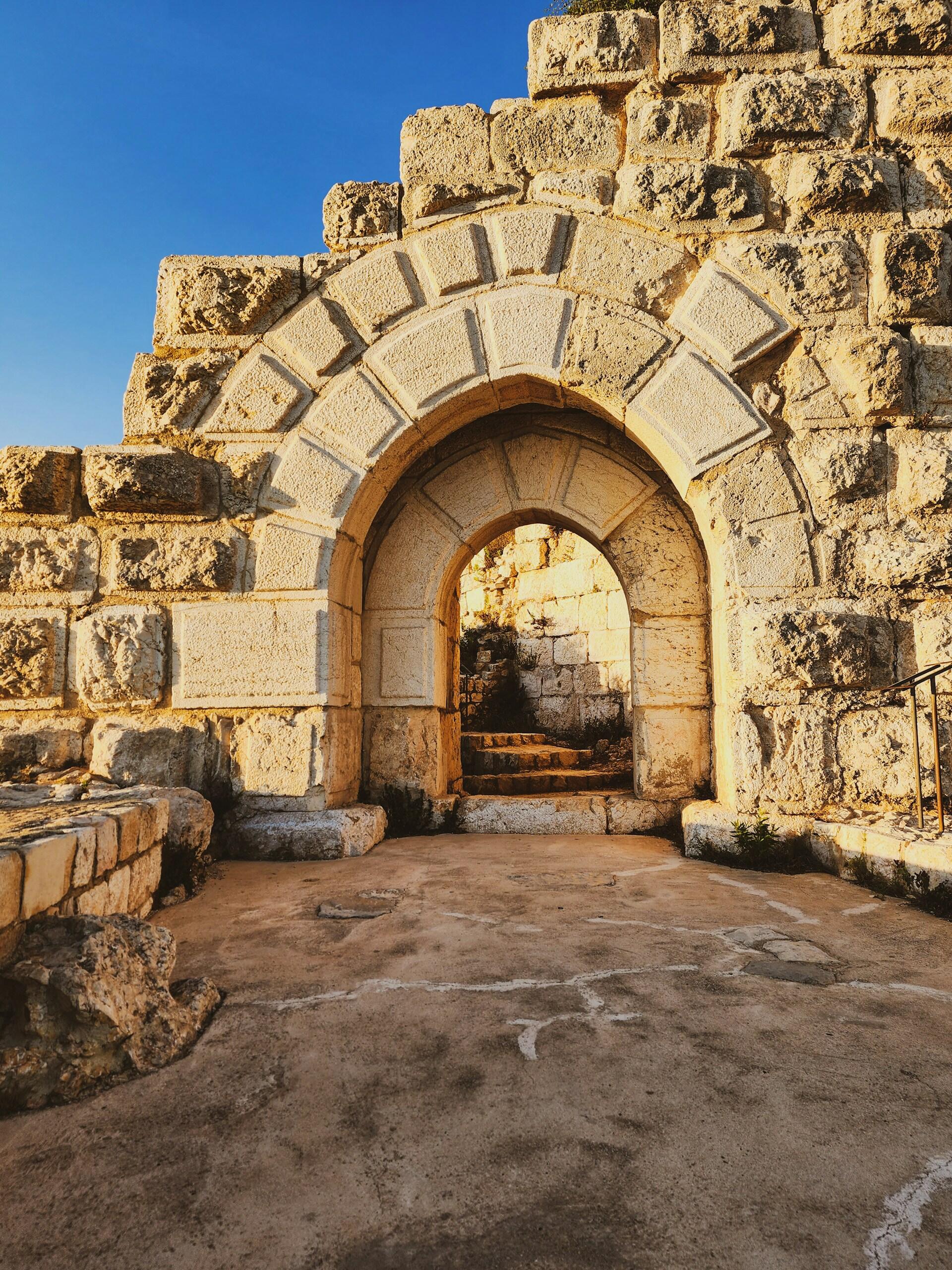 שער אבן עתיק בהריסות שטופות שמש, עם בנייה שחוקה, מדרגות ושמיים כחולים עזים