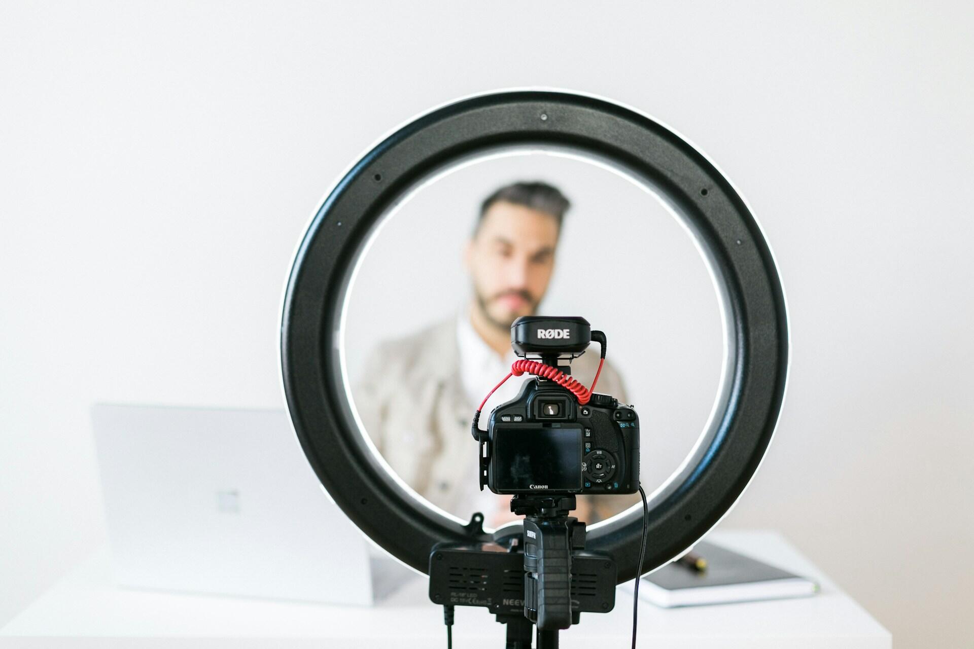 מצלמה על חצובה עם מיקרופון, ממוסגרת על ידי טבעת תאורה, על רקע לבן חלק ומחשב נייד בקרבת מקום