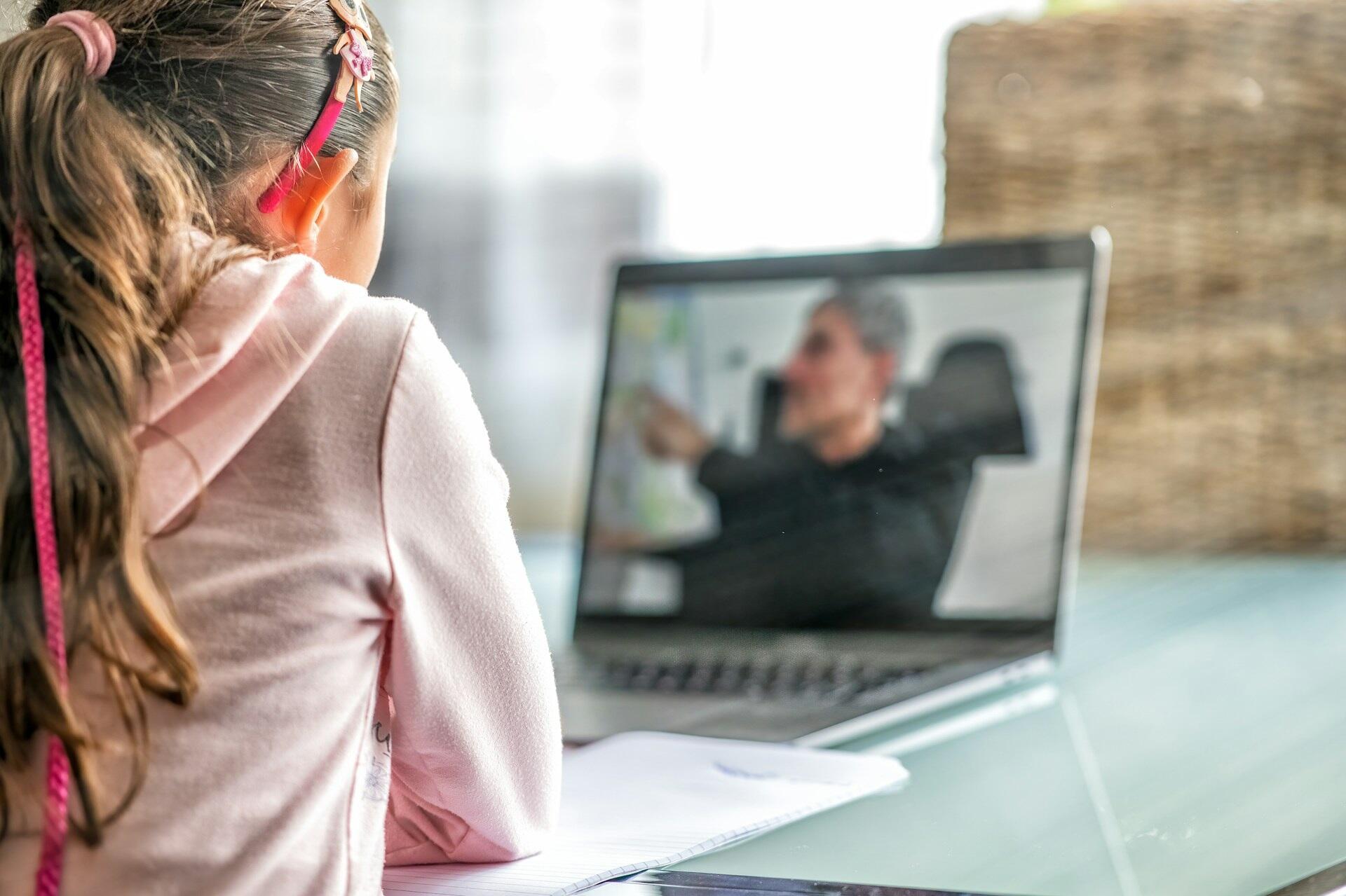 נערה צעירה עם קוקו יושבת ליד שולחן, צופה בתשומת לב במורה על מסך מחשב נייד במהלך שיעור וירטואלי