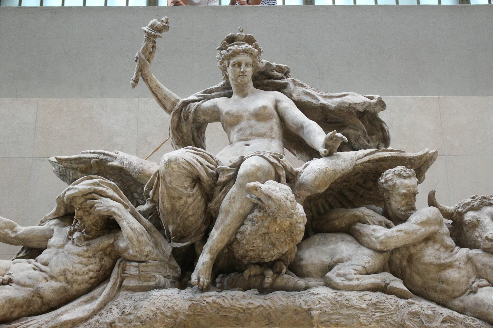 פסל מפורט מתאר דמות יושבת אוחזת חרב, מוקפת בדמויות אלגוריות של טבע ושלטון.