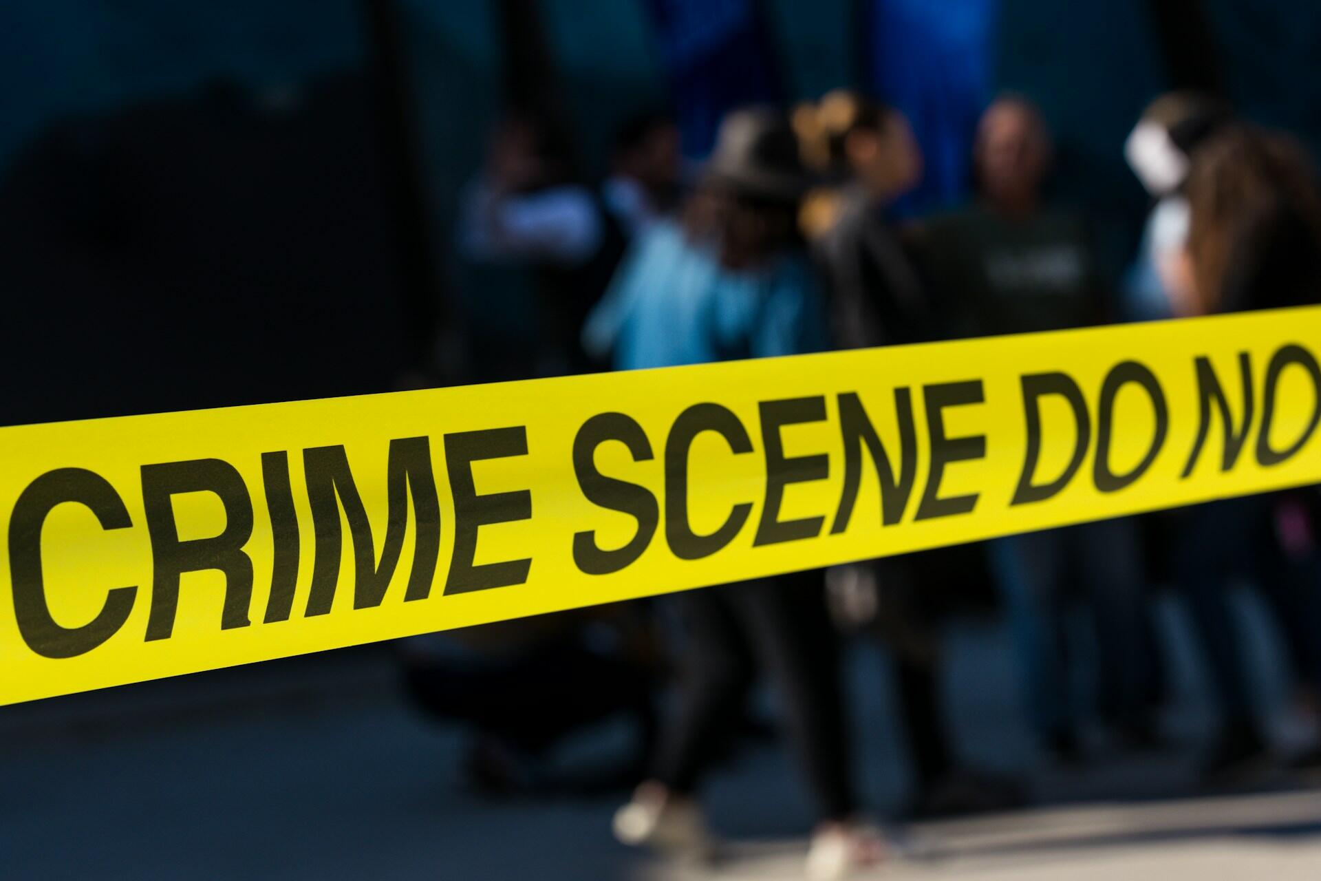 סרט זירת פשע צהוב בוהק עם הכיתוב "CRIME SCENE DO NO" נמתח בקדמת התמונה, כשברקע דמויות מטושטשות של אנשים