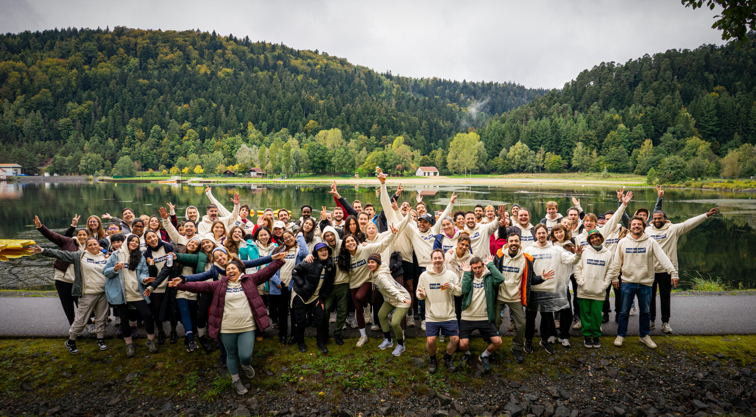 קבוצה גדולה של אנשים בבגדי חוץ קז'ואליים מצטלמים בשמחה ליד אגם עם גבעות מיוערות ברקע