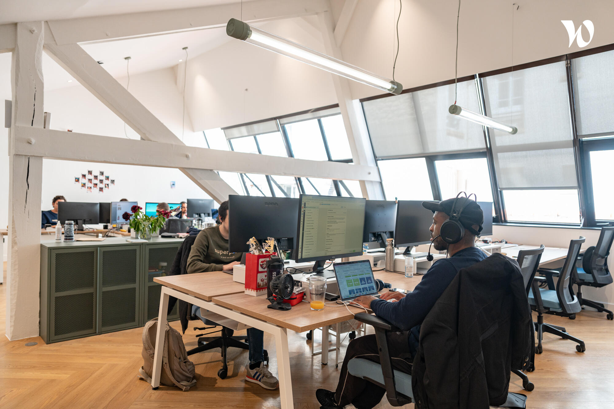 משרד פתוח מודרני עם עובדים העובדים ליד שולחנות עבודה המצוידים במחשבים, מחשבים ניידים ואוזניות תחת חלונות גדולים