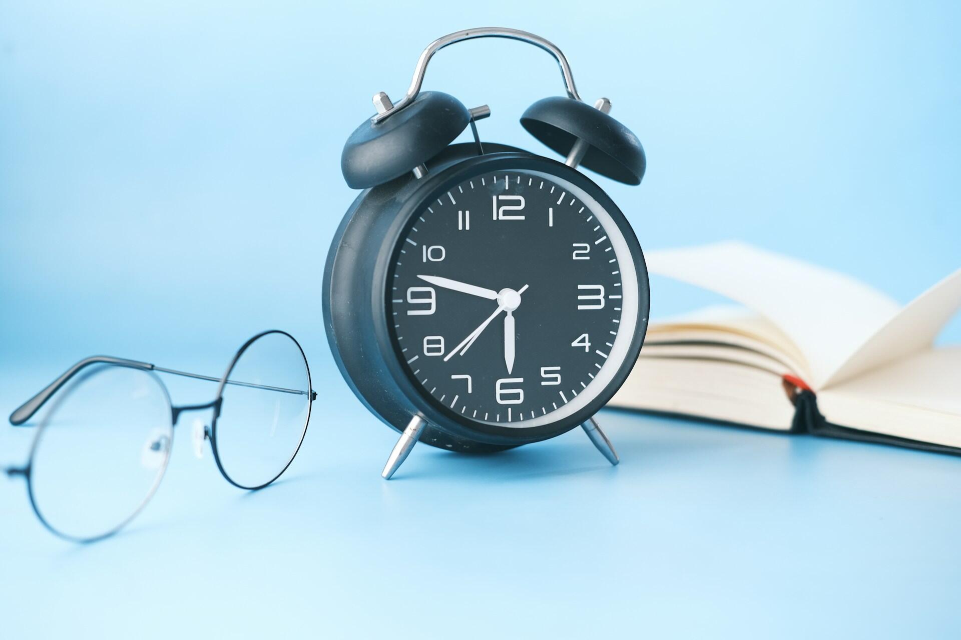 שעון מעורר רטרו שחור המציג את השעה 6:36 עם משקפיים וספר פתוח על משטח ורקע בצבע תכלת