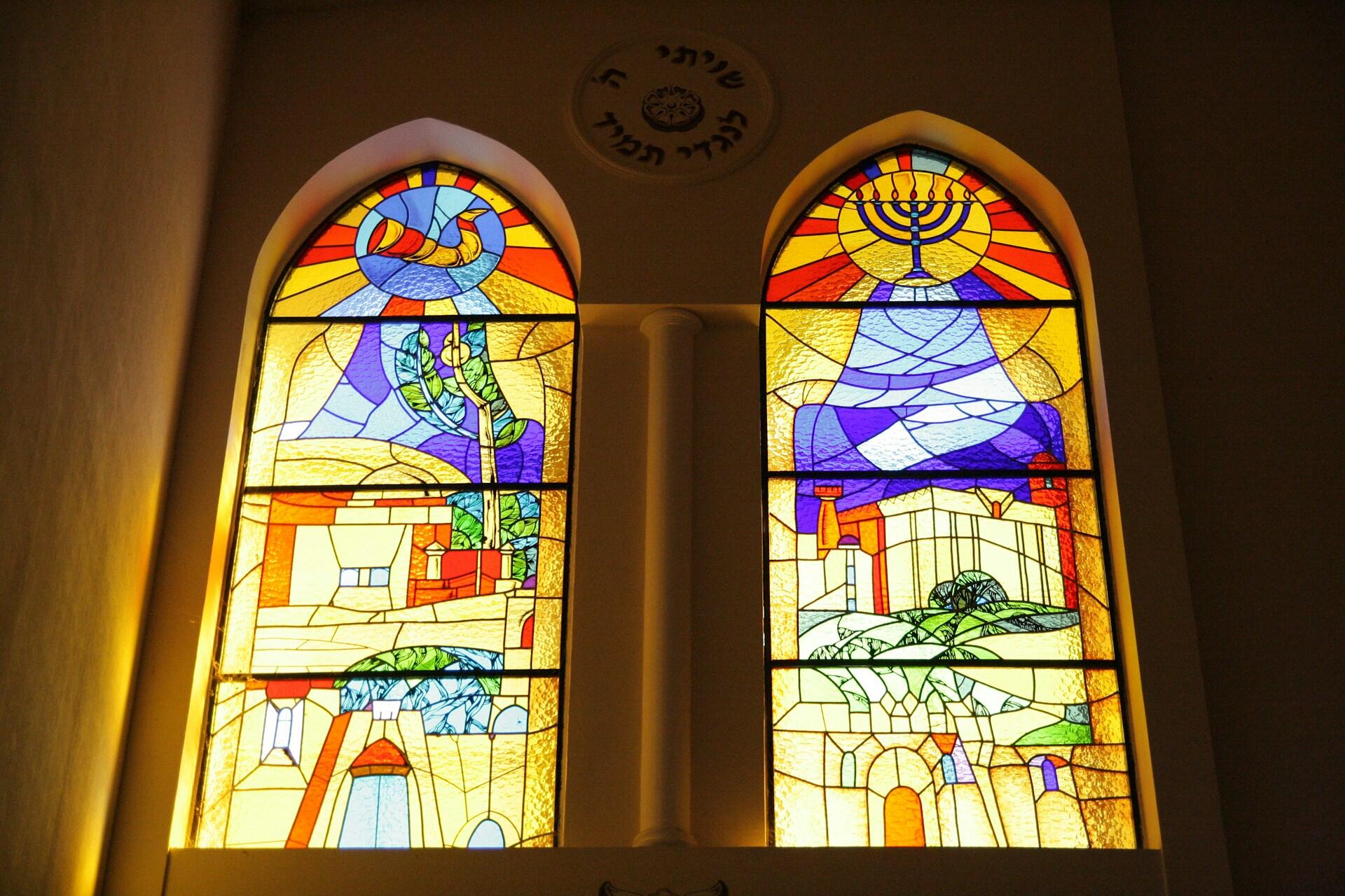 חלונות ויטראז' הכוללים שופר, חנוכייה ואלמנטים אדריכליים וטבעיים בגוונים עזים של צהוב, כחול, אדום וירוק