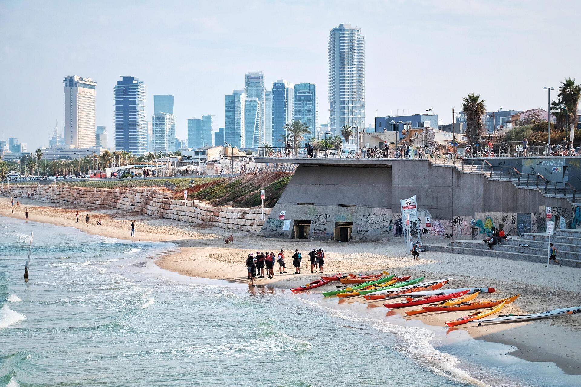 חוף ים עם קיאקים צבעוניים, אנשים התאספו על חוף חולי, קו רקיע מודרני של עיר ועצי דקל תחת שמיים בהירים