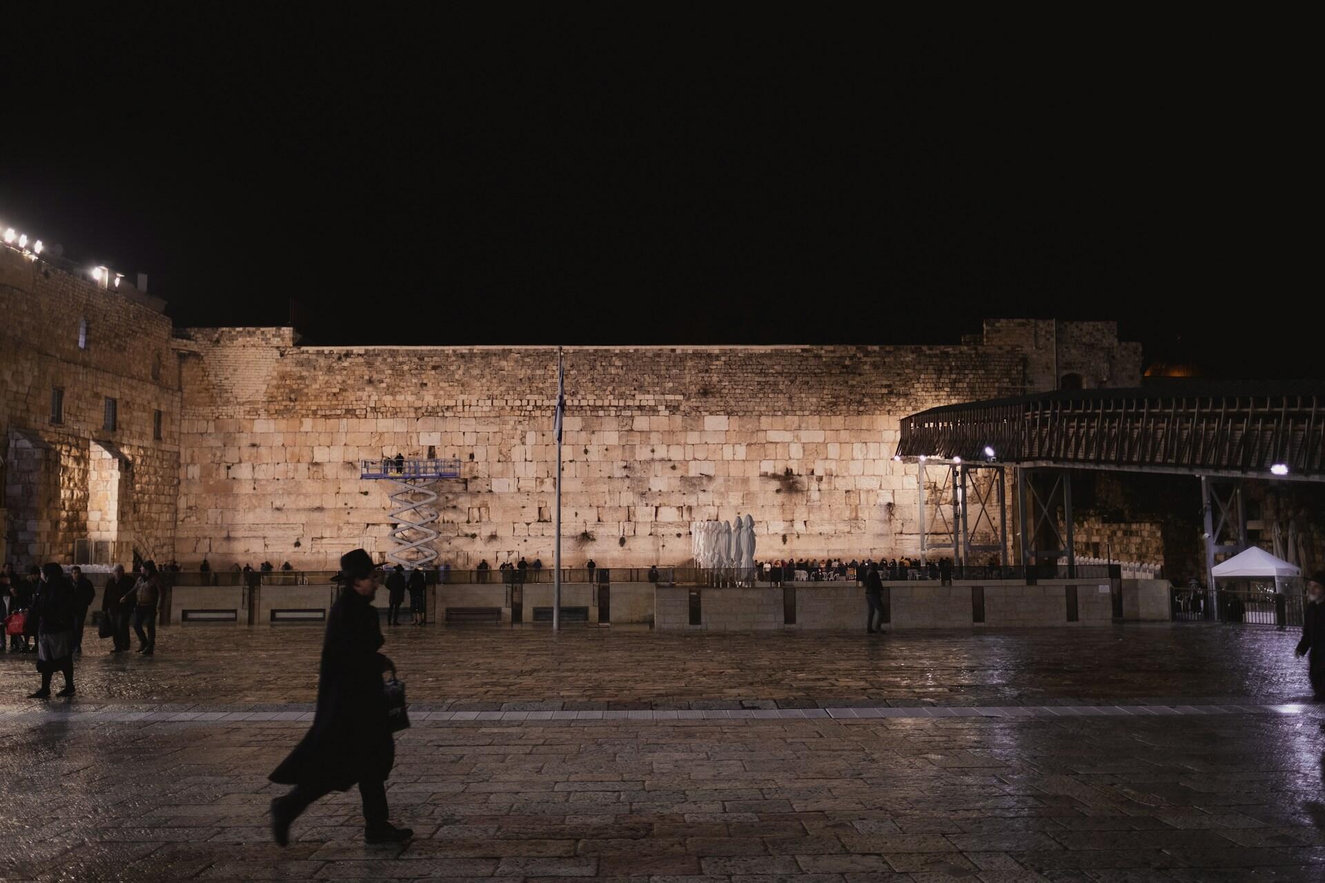 נוף לילי של רחבת הכותל המערבי עם אנשים הולכים ומתאספים, קיר אבן מואר ופלטפורמה מוגבהת בצד ימין