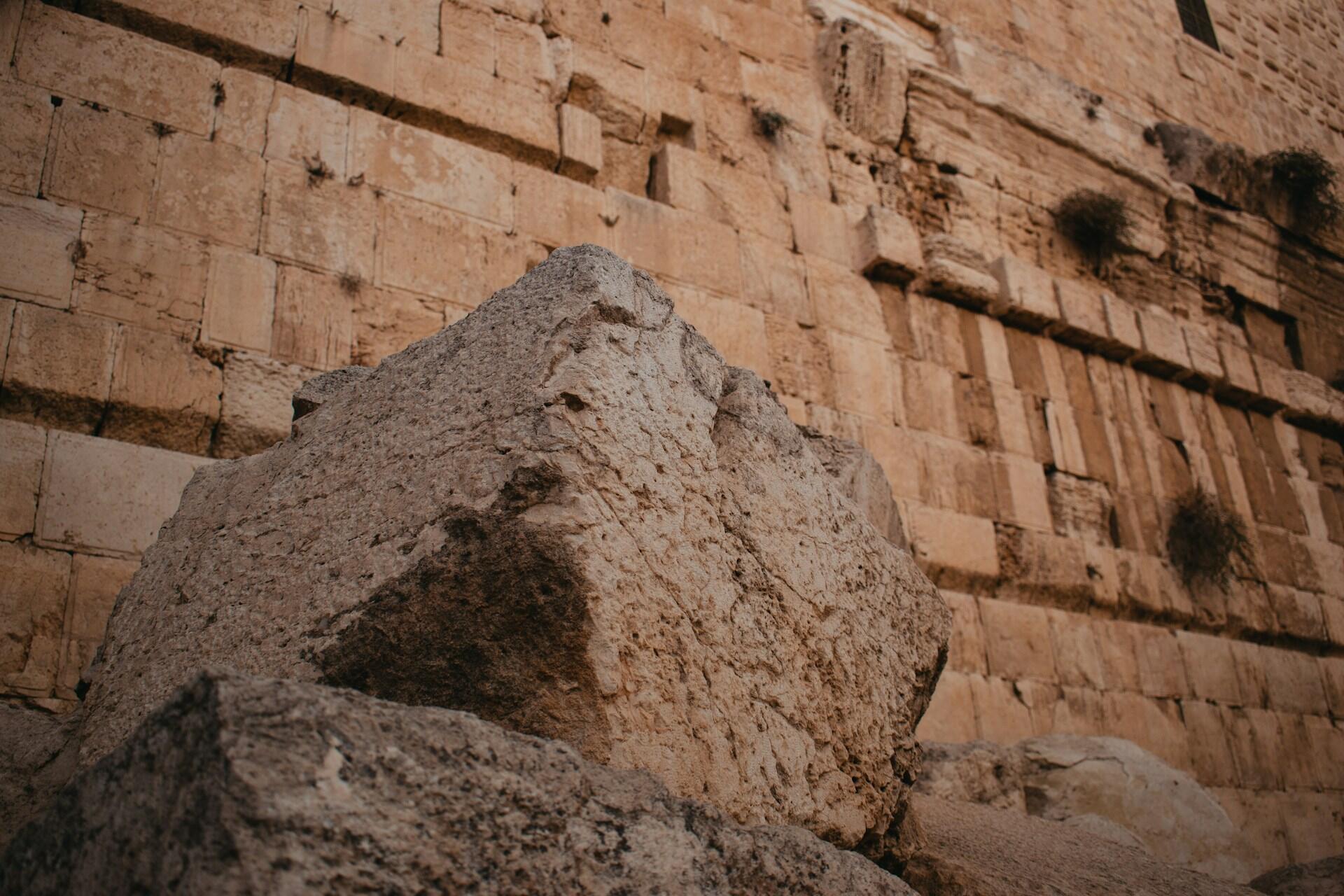 תקריב של גושי אבן עתיקים גדולים, בלויים, מוערמים ליד קיר לבנים בז' היסטורי עם צמחים קטנים הגדלים מתוך פערים