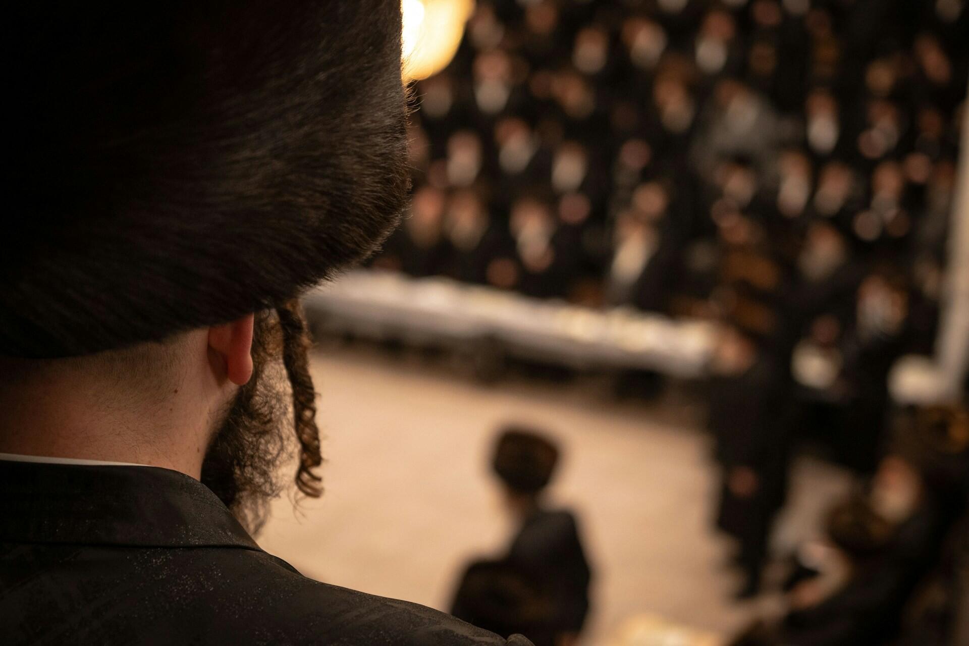 תקריב של גבר חובש כובע פרווה מסורתי עם מנעולי צד, צופה ברקע בהתכנסות מטושטשת של אנשים בלבוש דומה
