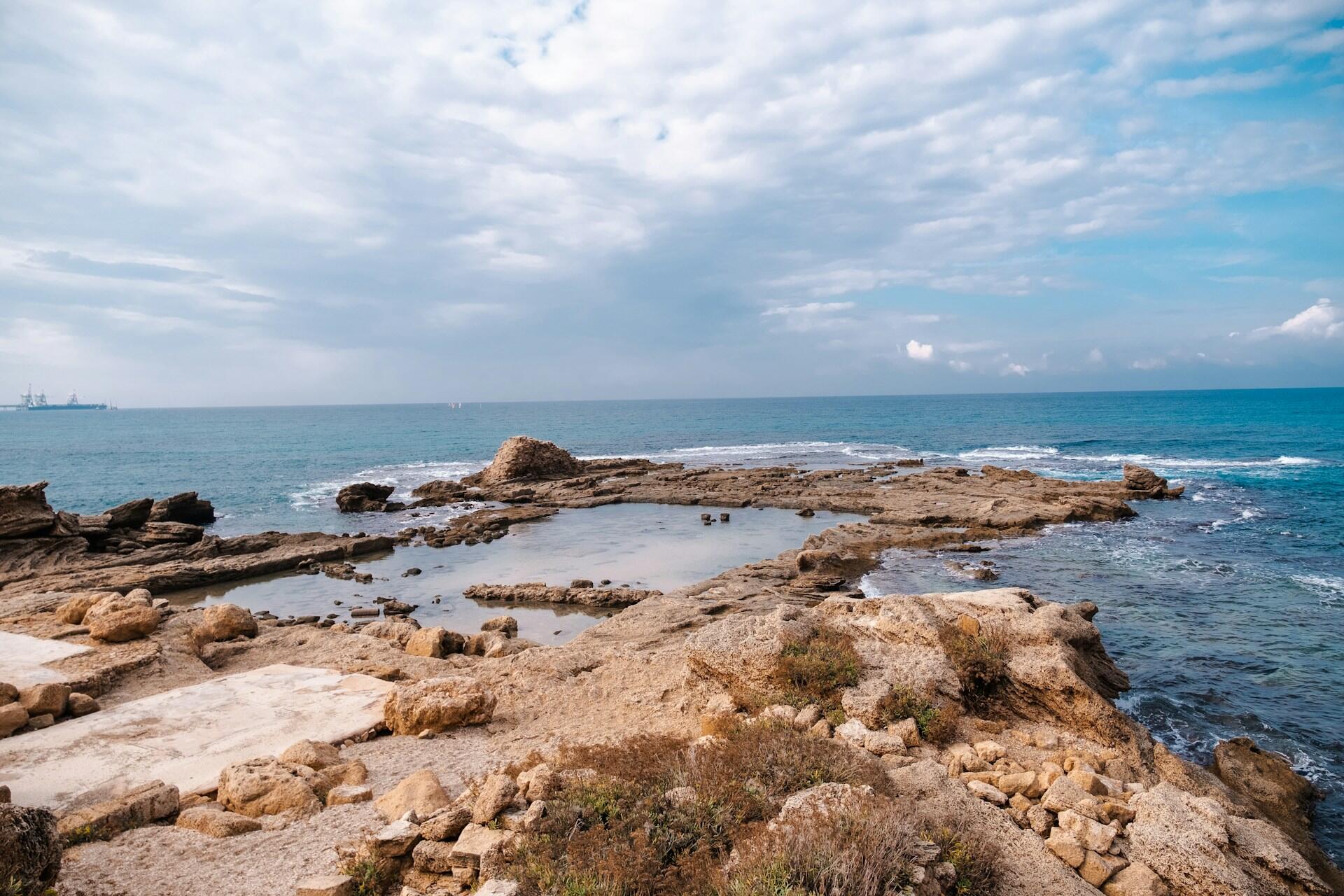 קו חוף ים תיכוני סלעי ובו בריכות גאות ושפל ותצורות אבן מחוספסות תחת שמיים כחולים מעוננים, כשספינות נראות באופק הרחוק