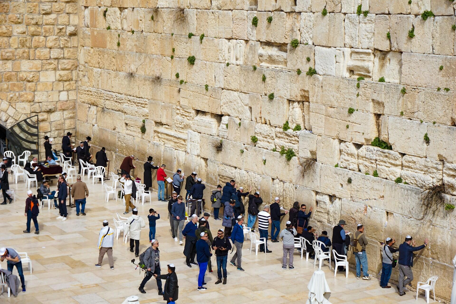 אנשים התאספו והתפללו ליד הכותל המערבי, מבנה אבן גיר היסטורי בירושלים, חלקם עומדים ואחרים יושבים על כיסאות לבנים