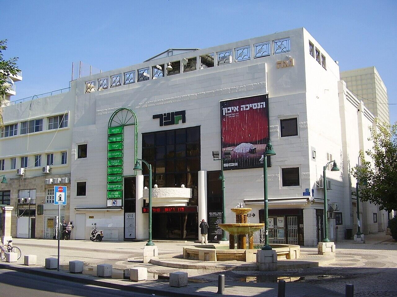 בניין מודרני לבן בן מספר קומות עם חלונות גדולים, מזרקה מרכזית, פנסי רחוב ירוקים ושילוט בעברית תחת שמיים כחולים וצלולים