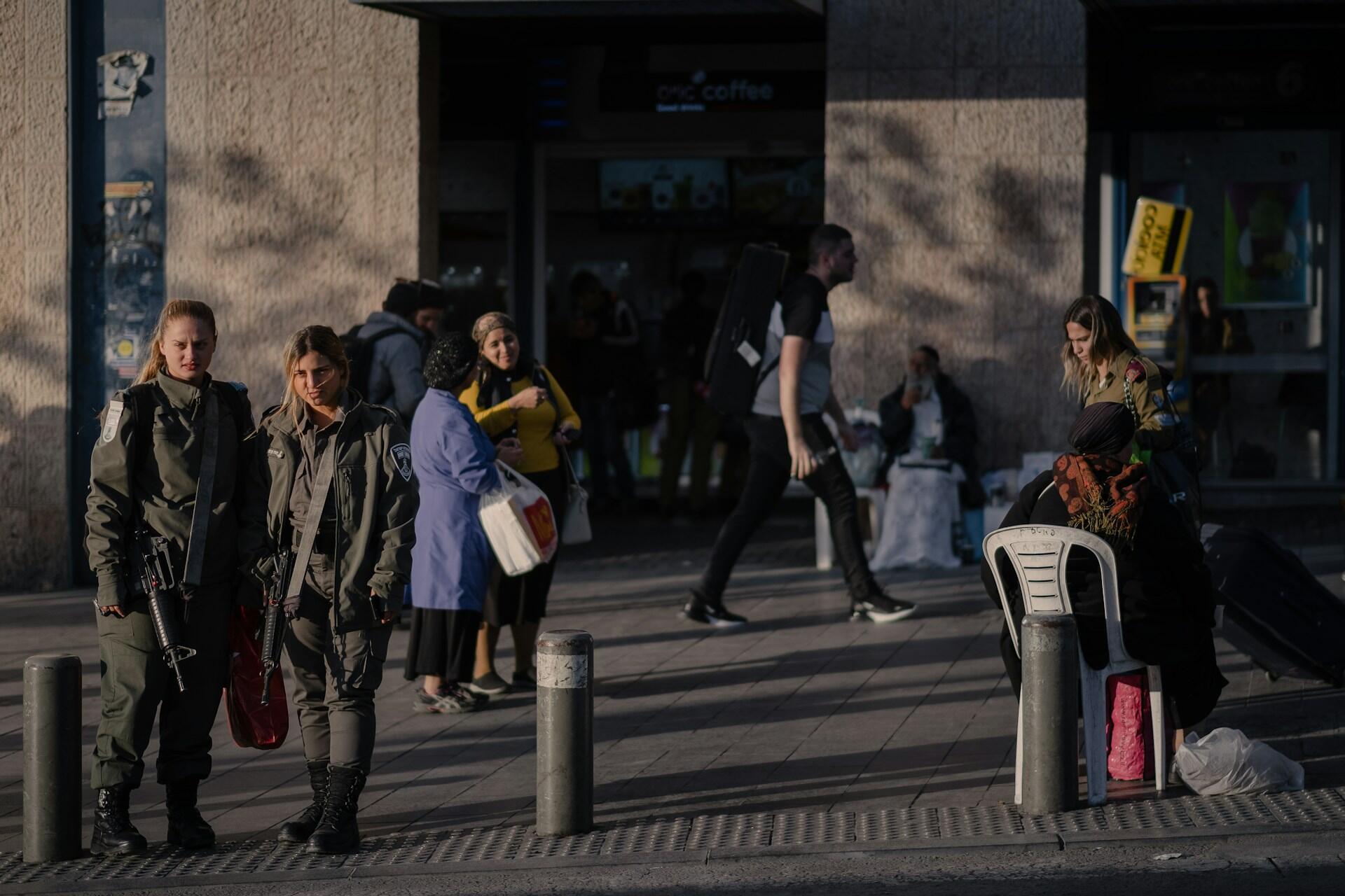 שני שוטרים חמושים עומדים על מדרכה סואנת כשבקרבתם הולכי רגל נמצא אדם יושב עם מזוודות