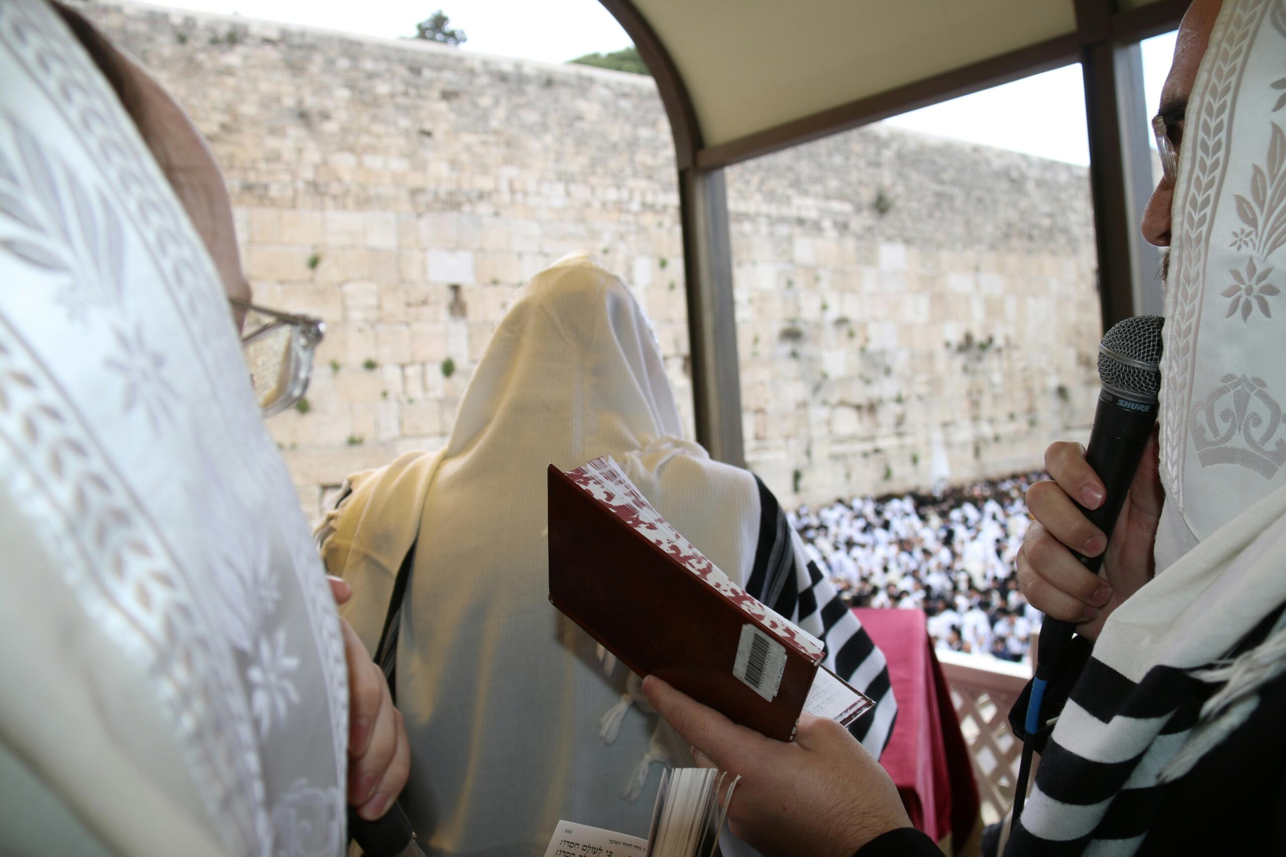 גברים בטליתות יהודיות מסורתיות מתאספים בכותל המערבי, אחד אוחז בסידור תפילה ואחר מדבר לתוך מיקרופון