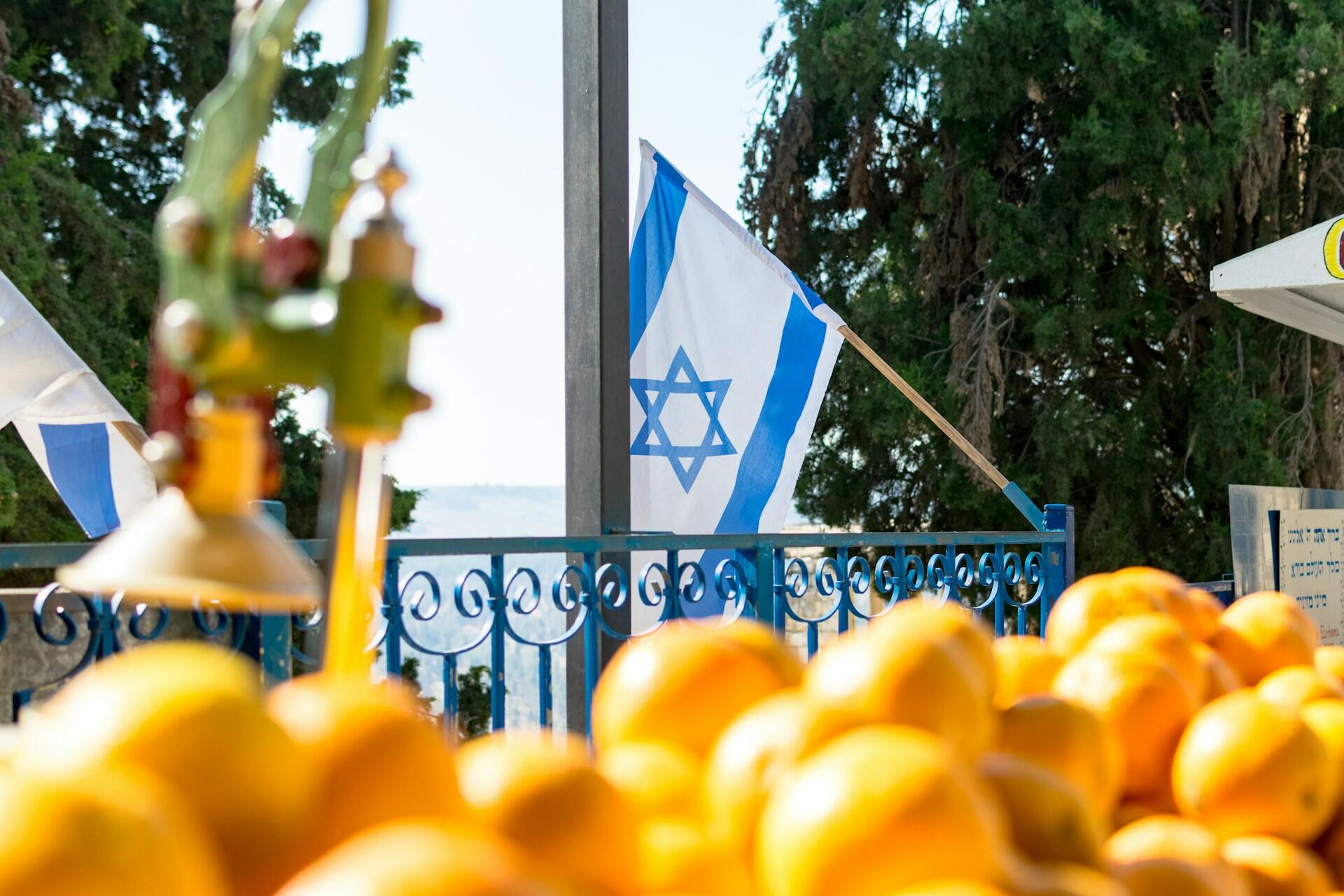 פירות כתומים בחזית עם דגל ישראל ועצים ברקע, נצפים מבעד למעקה מתכת כחול