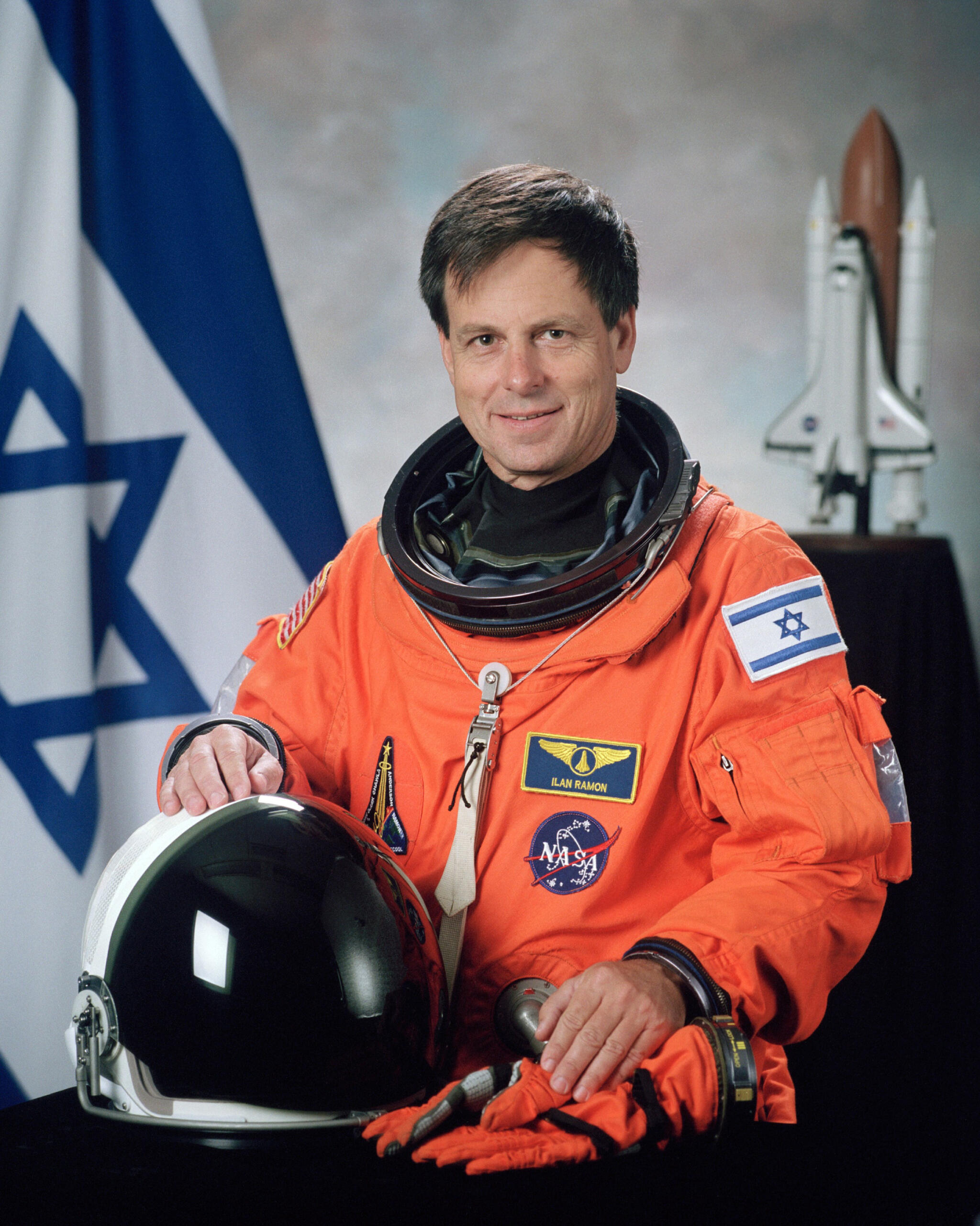 האסטרונאוט הישראלי אילן רמון בחליפת חלל כתומה, אוחז בקסדתו לצד פנים של חללית