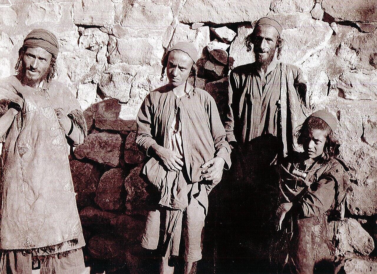 תצלום בשחור-לבן של ארבעה אנשים בלבוש מסורתי, העומדים על רקע קיר אבן גס