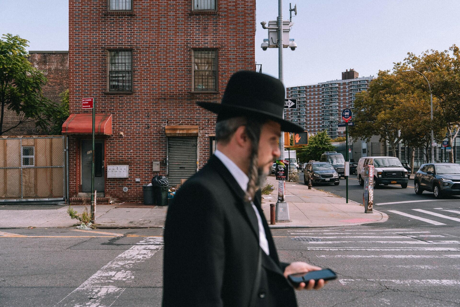 גבר בחליפה וכובע שחורים בודק את הטלפון שלו בעודו חוצה רחוב עירוני סואן