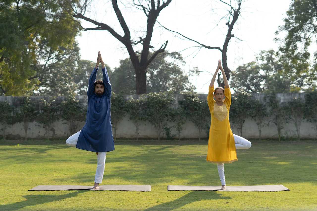 A man and a women practice tree pose Vriksasana while wearing kurtas and churidar.