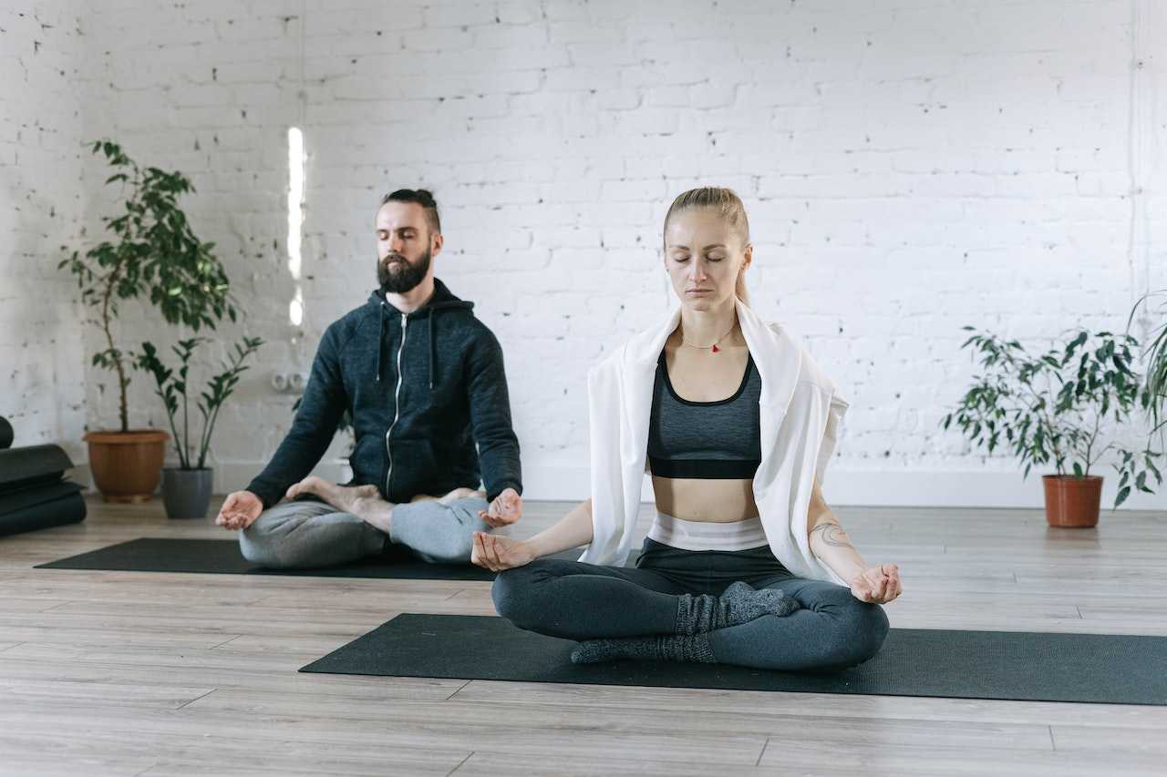 A man and a woman sit on yoga mats and breathe. The man is wearing a zipped hoodie and lounge pants and the woman is wearing a sports top under an unzipped hoodie and yoga pants.