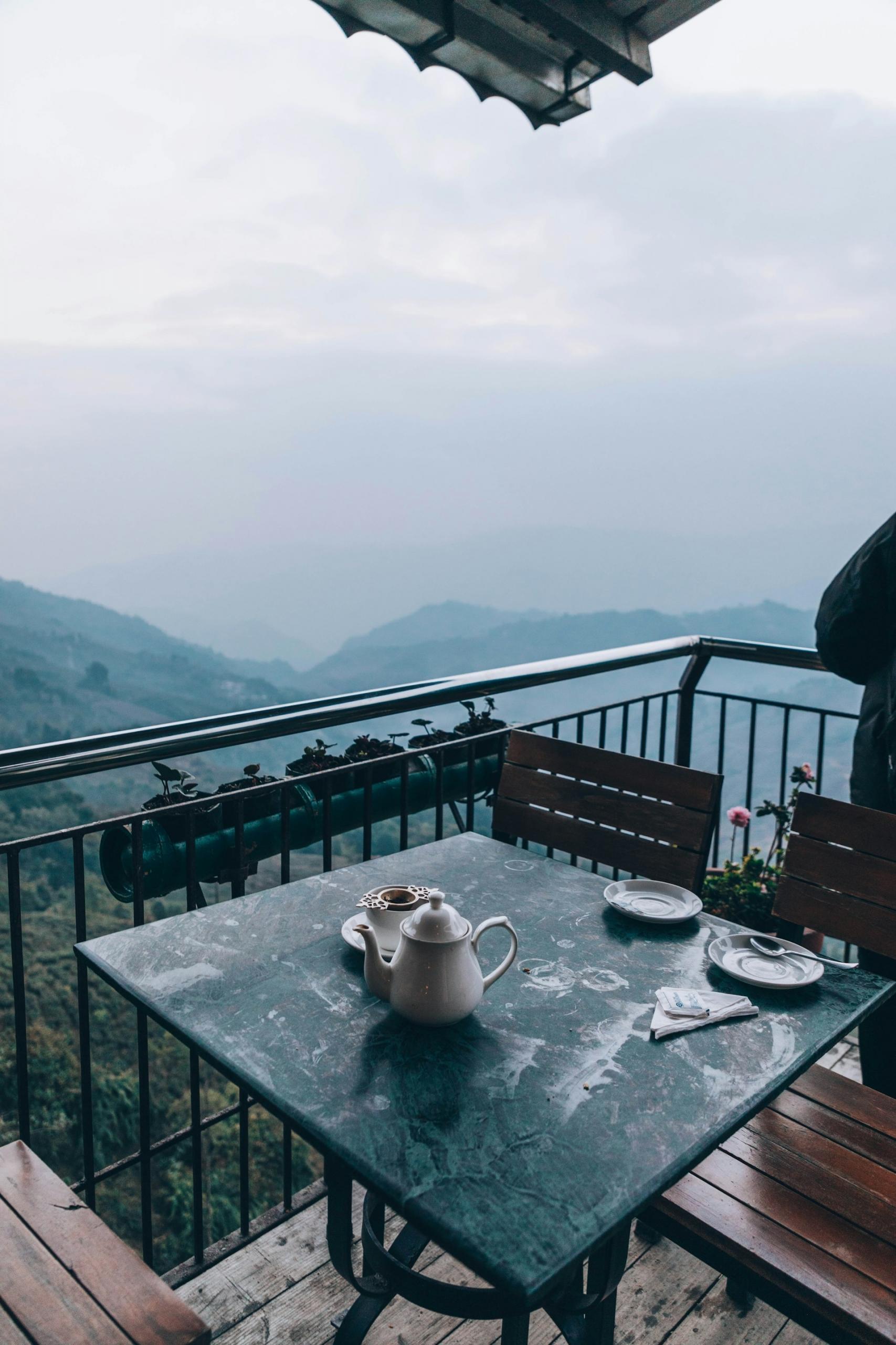 A cafe table with a teapot and tea cups on a veranda overlooking the misty forests of Darjeeling