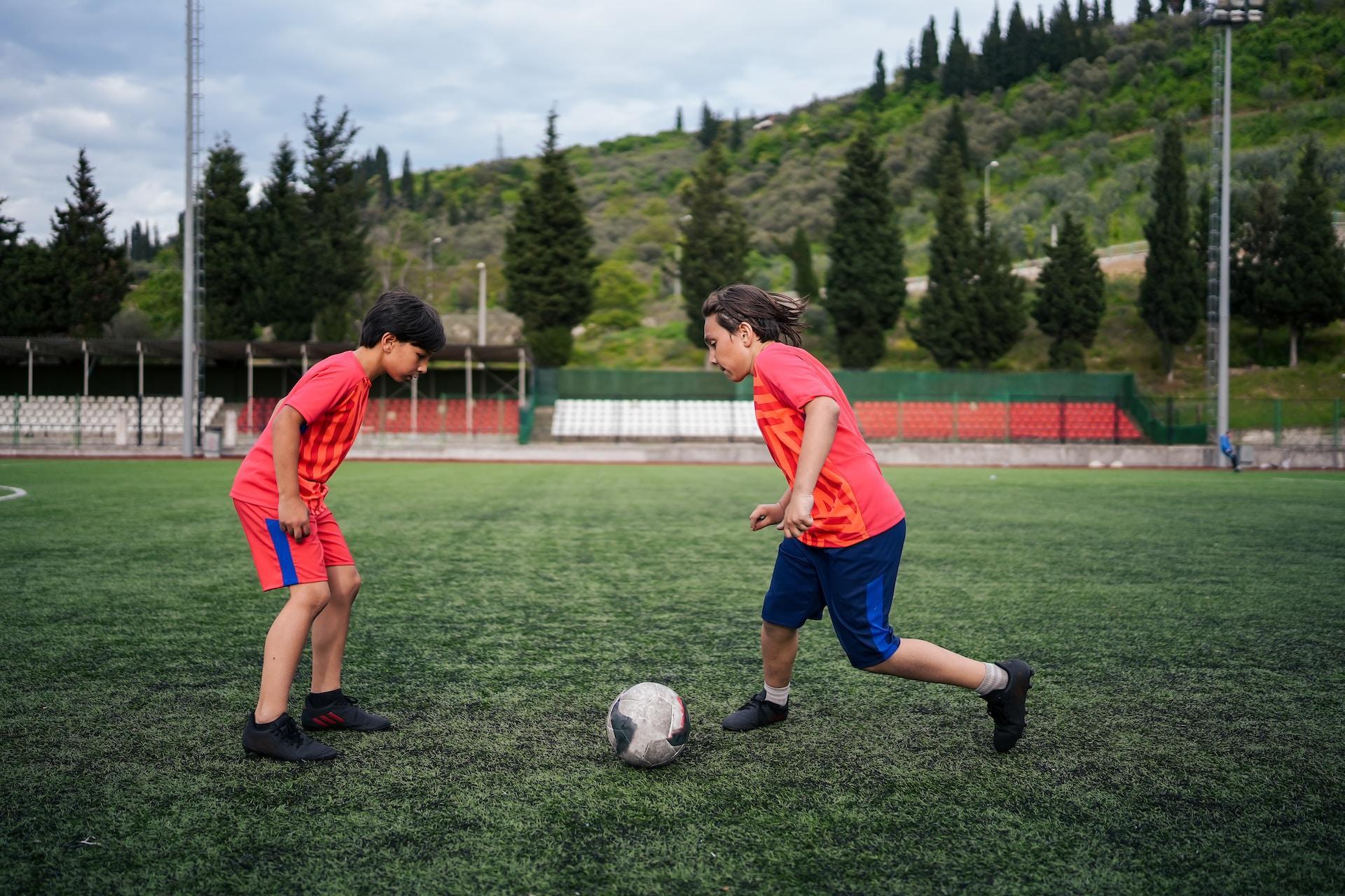 Two youths in orange jerseys, one wearing blue shorts and the other red, face off over a soccer ball in an otherwise empty pitch.