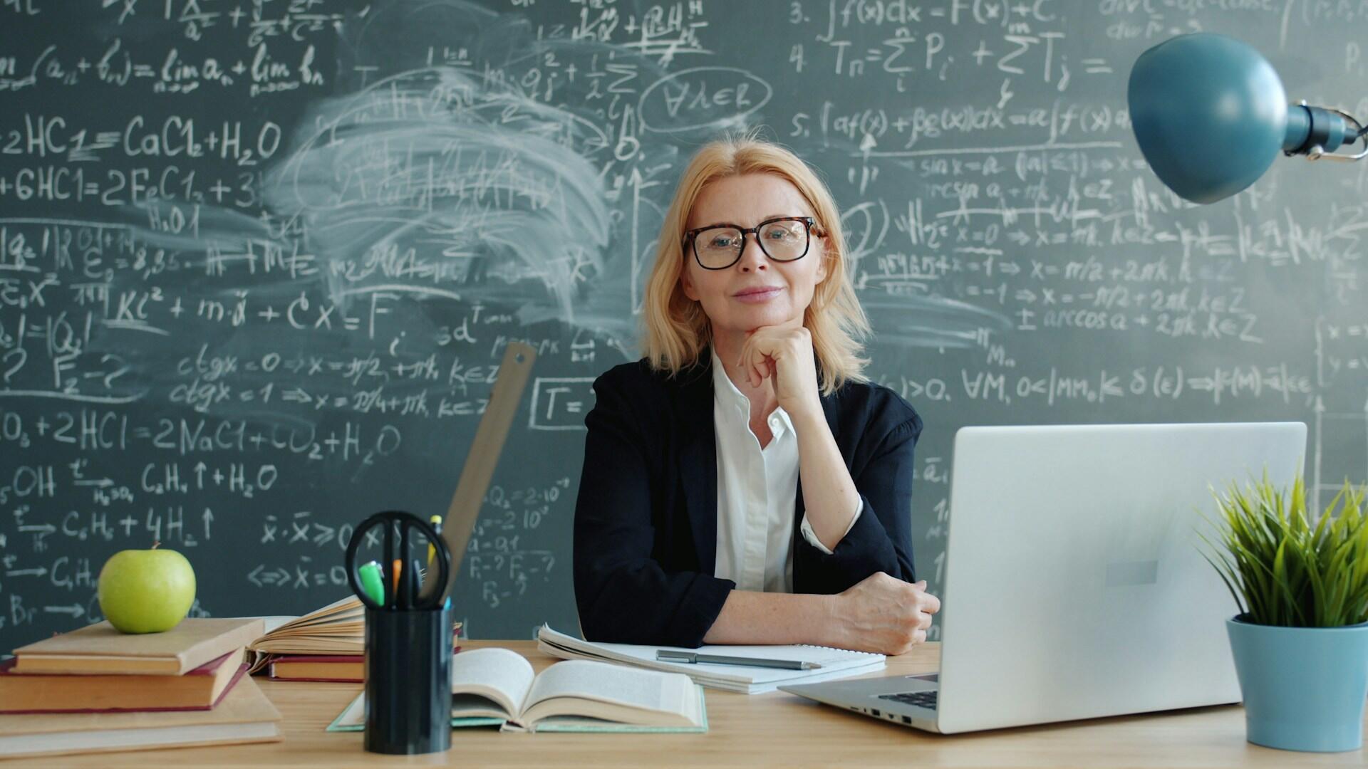 Professoressa universitaria seduta alla scrivania davanti a una lavagna con formule.