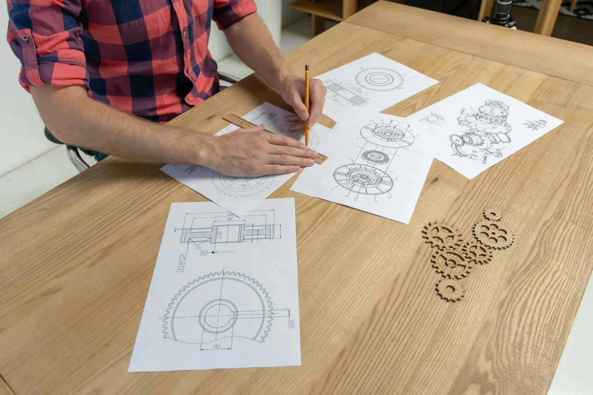 Una persona seduta a un tavolo di legno mentre disegna e misura componenti meccanici su fogli tecnici. Intorno a lui ci sono schemi di ingranaggi e pezzi meccanici, oltre a piccoli modelli di ruote dentate in legno.