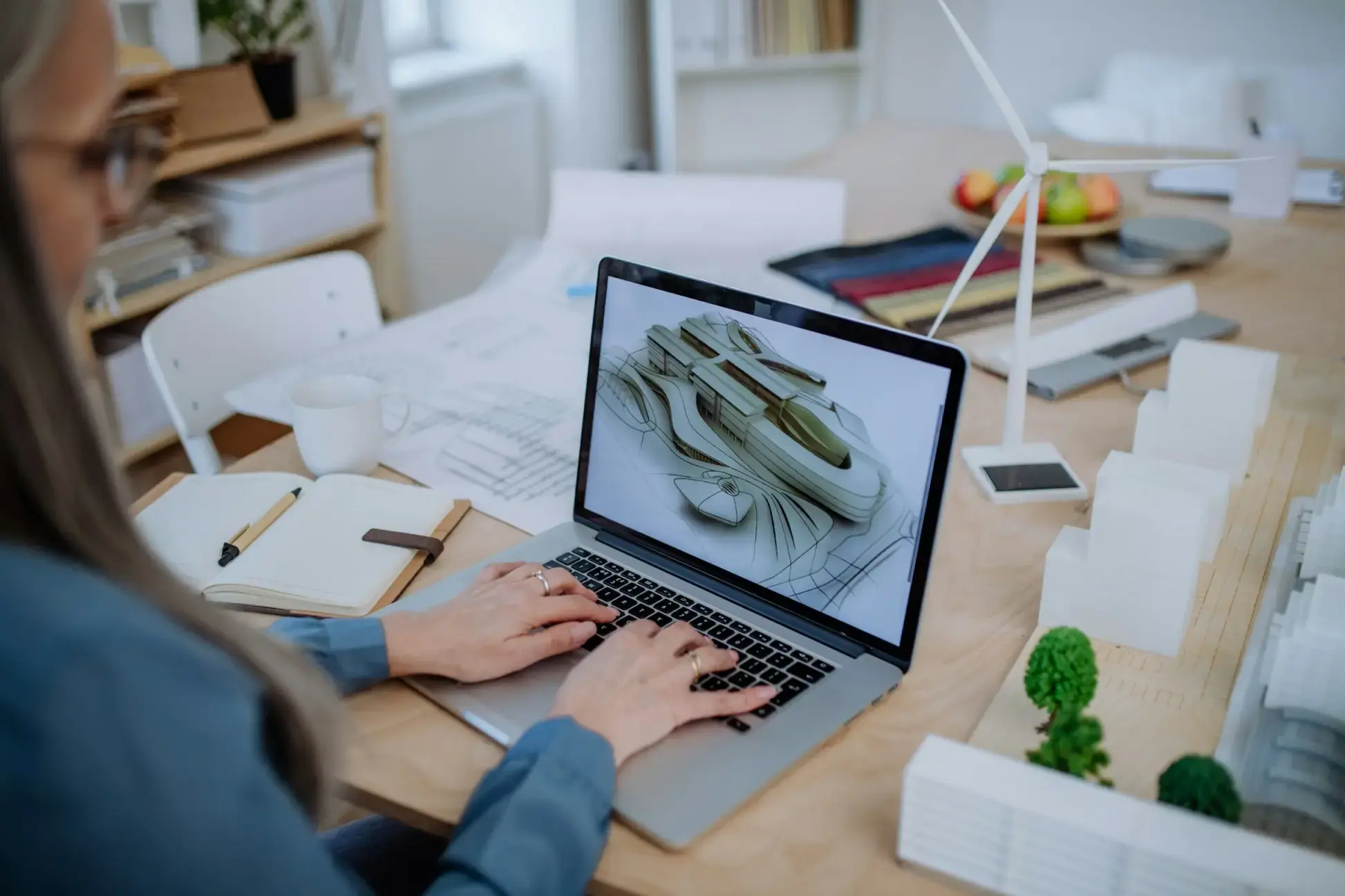 Una persona che lavora al computer su un modello 3D, circondata da schizzi, un quaderno e materiali di progettazione su un tavolo.