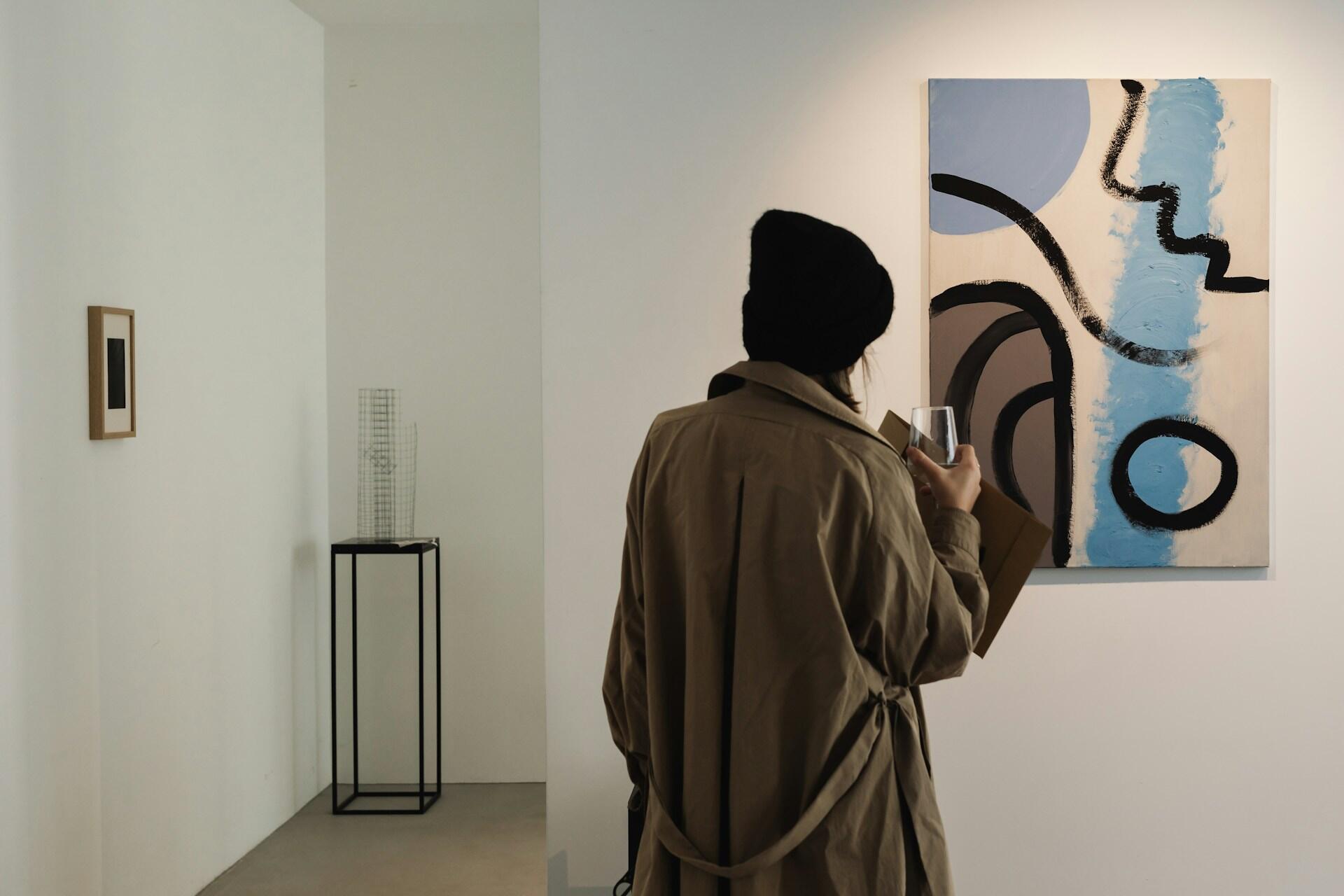 Una ragazza osserva un quadro contemporaneo mentre sorseggia un bicchiere di vino. 