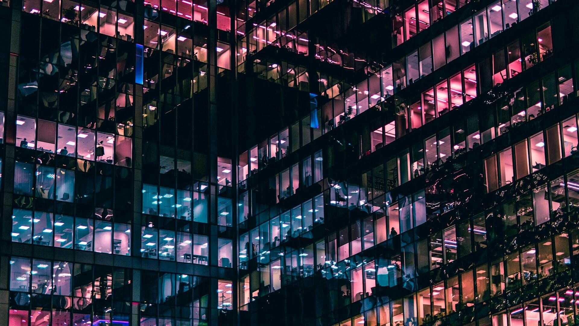 Un palazzo di vetro con dentro uffici illuminato da luci viola e blu. 