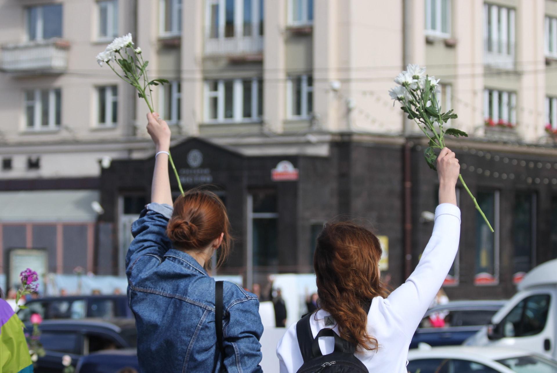 Deux jeunes femmes de dos dans la rue, un bras levé, tenant chacune une grande fleur