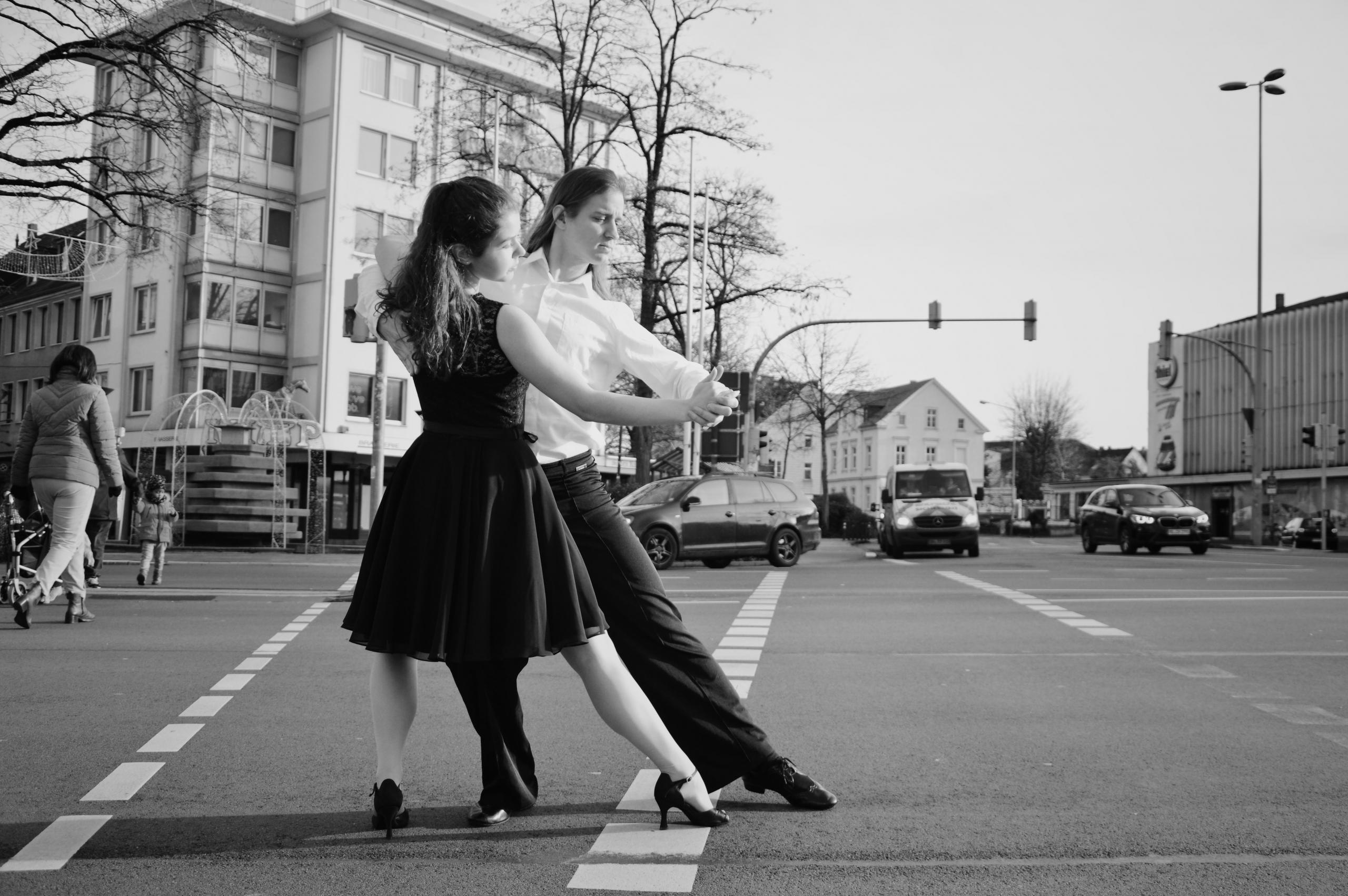 Couple qui danse le tango dans un décor urbain, sur la chaussée, au milieu de quelques voitures et piétons, noir et blanc.