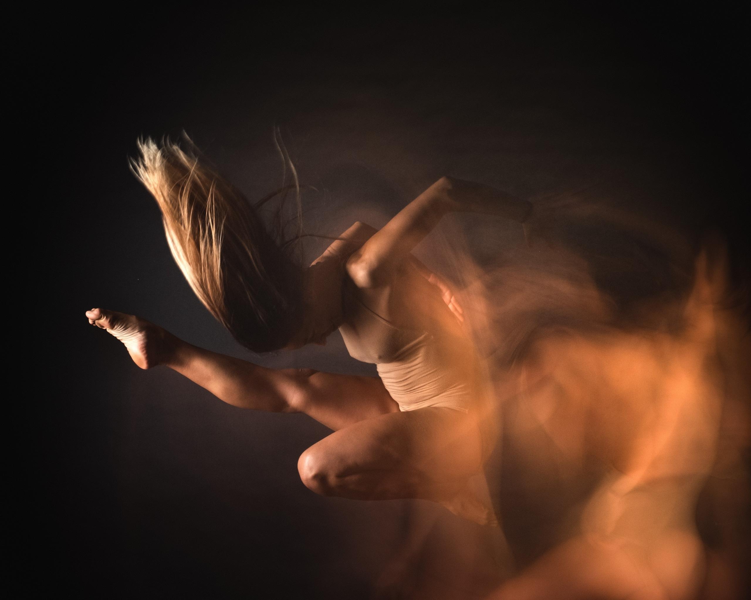 Une photo d'une danseuse en train de faire une figure de classique dans les airs en lançant du sable. Fond noir, spot de lumière avec des reflets or.