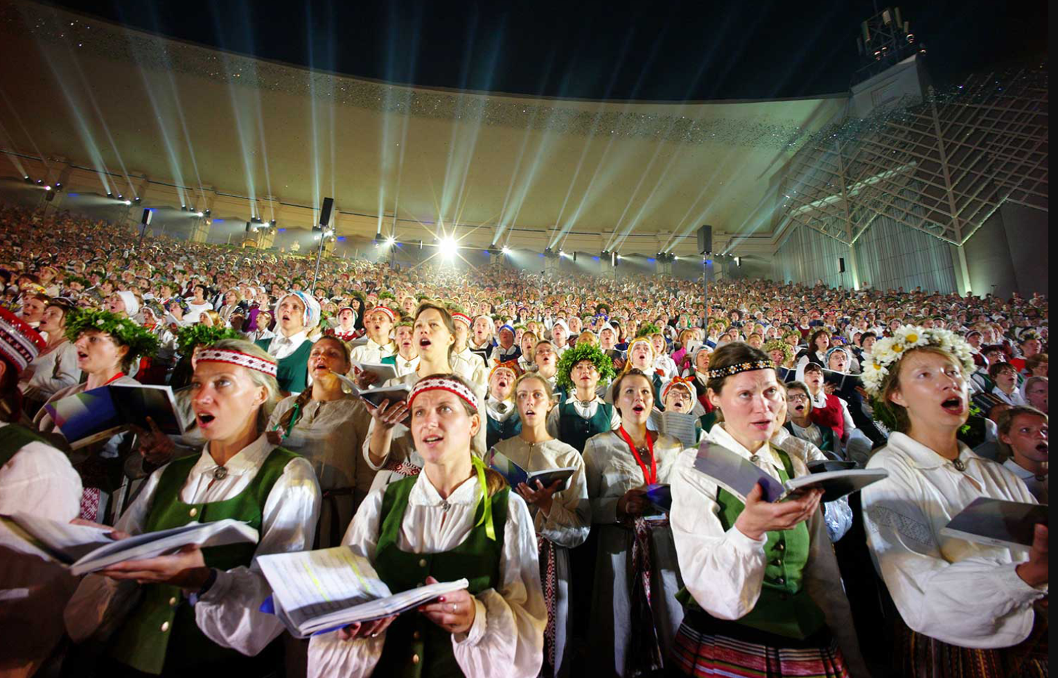 Liels tautastērpos tērpts koris dzied plašā, spoži apgaismotā estrādē.