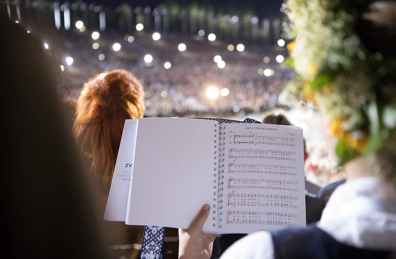 Dziedātājs tur atvērtu nošu grāmatu lielā, izgaismotā koru koncertā.