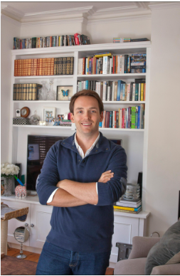 Marks Makleins stāv pie grāmatu plaukta.