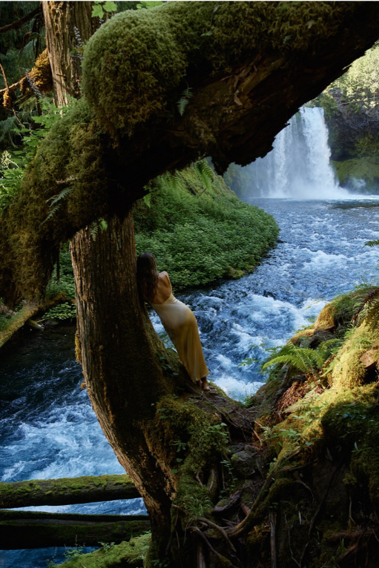 Sieviete stāv uz sūnaina koka saknēm pie upes.