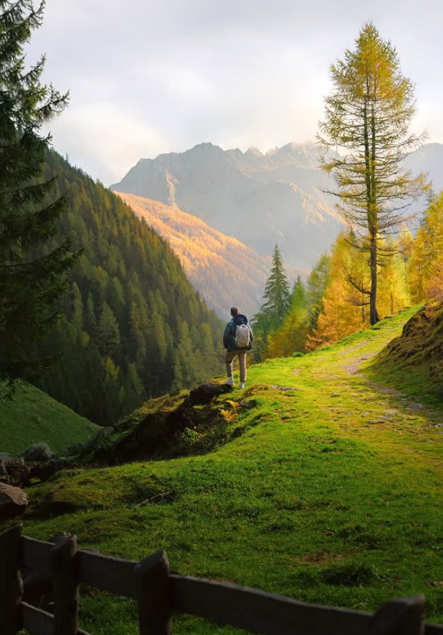 Cilvēks ar mugursomu stāv uz zaļa kalnu takas nogāzes.