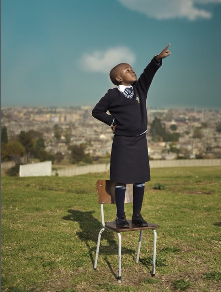 Skolniece stāv uz krēsla brīvā dabā  ar paceltu roku.