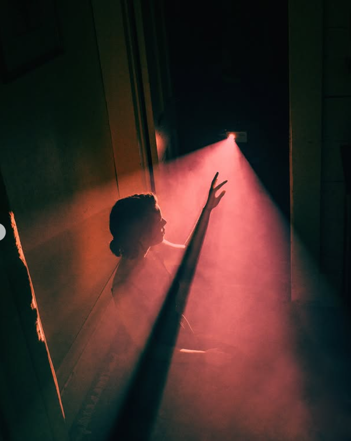 Sievietes siluets tumšā telpā sniedzas pret rozā gaismas staru.