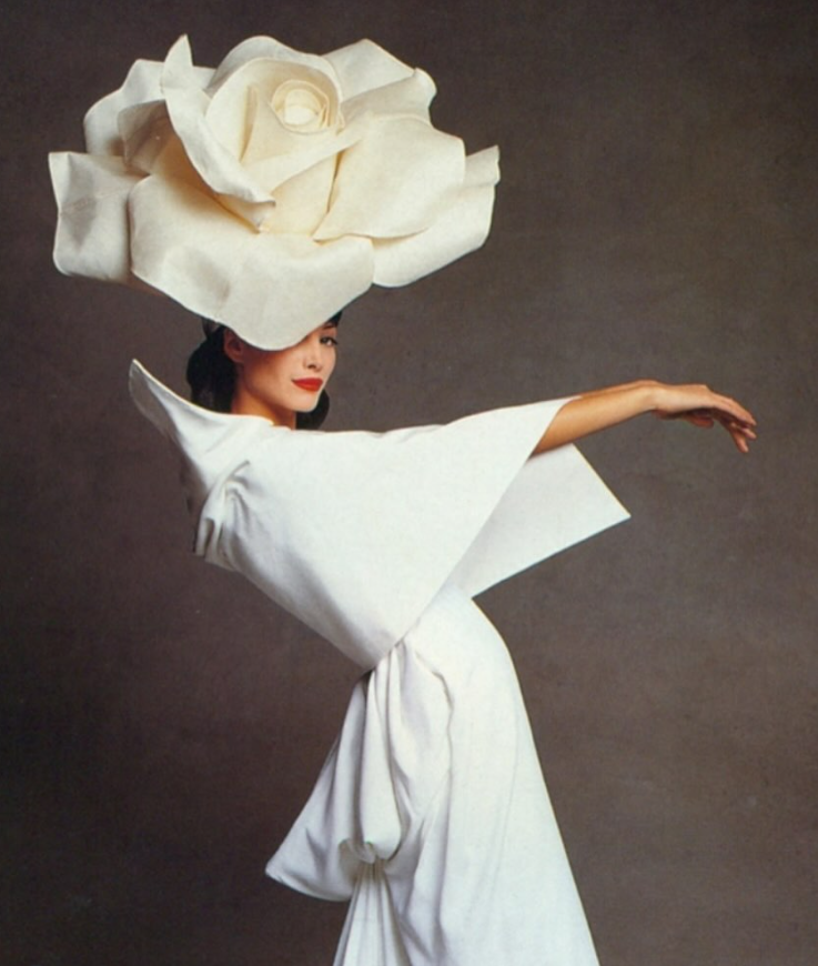 Sieviete baltā tērpā ar lielizmēra zieda galvassegu izstieptā, elegantā pozā.