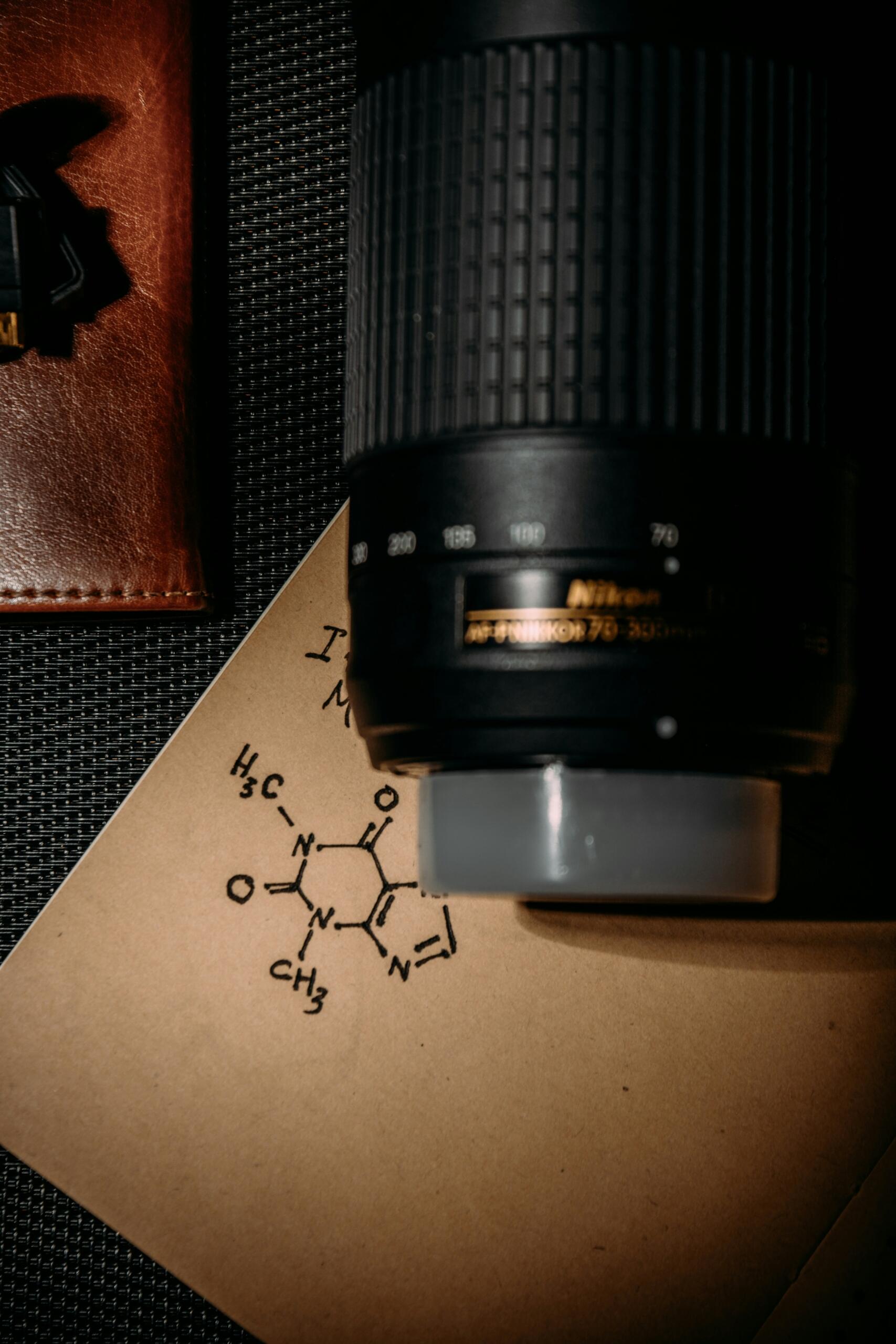 Objektīvs blakus piezīmēm ar uzzīmētu ķīmiskā savienojuma formulu uz papīra.