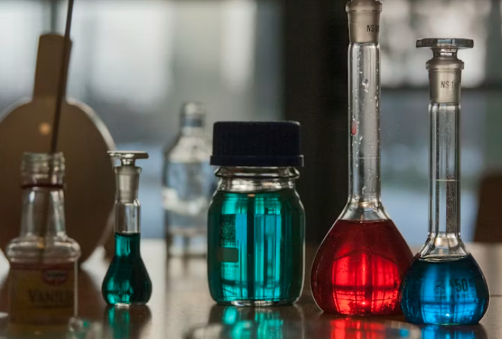 Laboratorijas trauki ar krāsainiem šķīdumiem, izvietoti uz galda ķīmijas laboratorijas vidē.